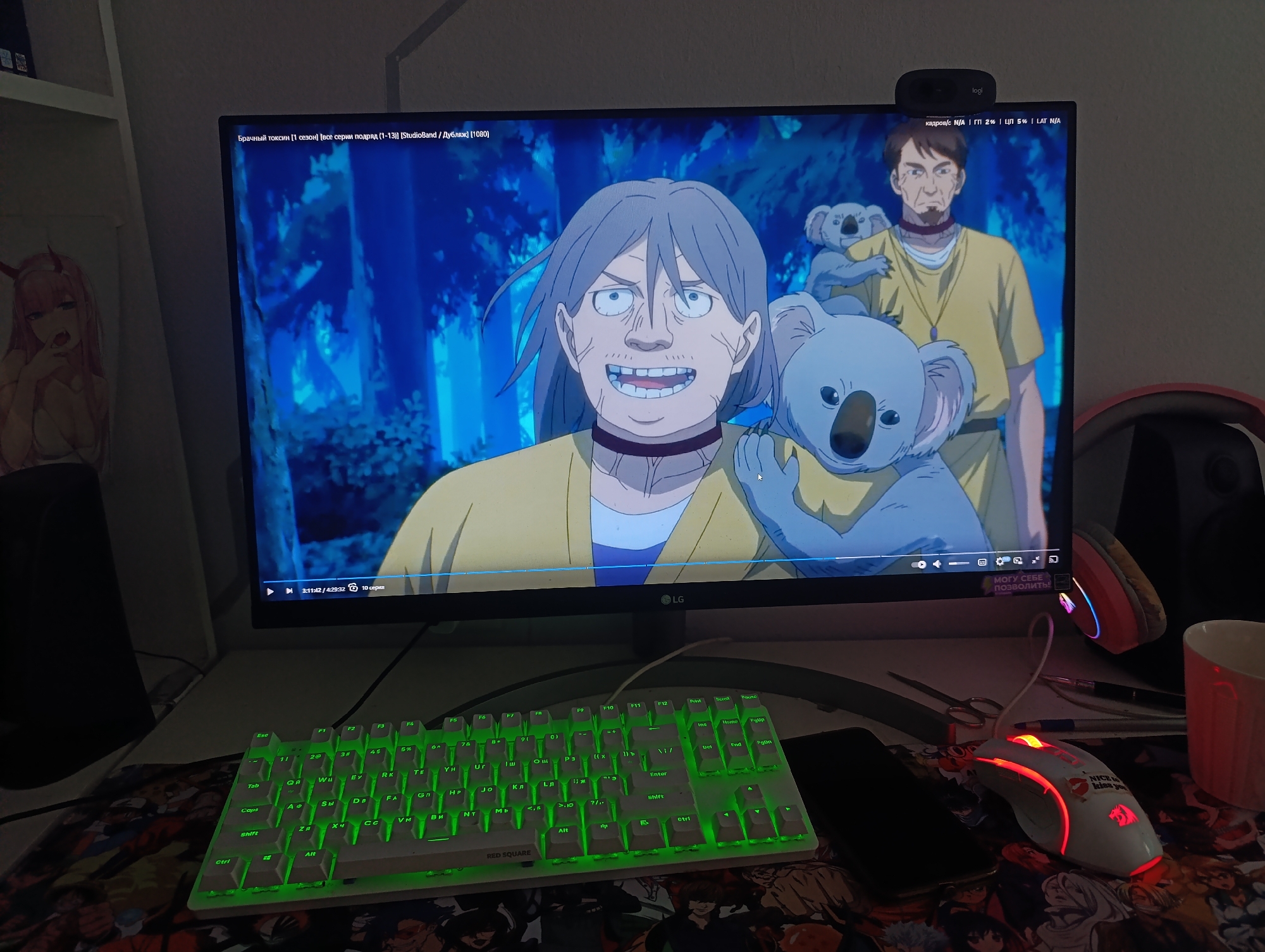 The image shows a computer monitor displaying an anime scene. On the monitor, two characters and two koalas are visible. The character in the foreground is a man with gray hair, wearing a yellow shirt and a choker. He has a wide smile and appears to be looking directly at the viewer. Behind him, another man in a yellow shirt with dark hair is visible, holding a koala on his shoulder. In front of this man, a larger koala is held by the foreground character. The background of the anime scene is a forest with blue and green tones.
Below the monitor, a white keyboard with green backlighting is visible. To the right of the keyboard, a white computer mouse with red lighting is present. The mouse has a slogan on it: "NICE TO KISS YOU". A dark-colored smartphone is located to the left of the mouse. The scene appears to be a desk setup in a room.
The provided information states the location is the city of Abakan, Russia. However, there are no visual cues in the image that specifically identify it as being in Abakan. The image depicts an indoor setting with common household items and electronics.