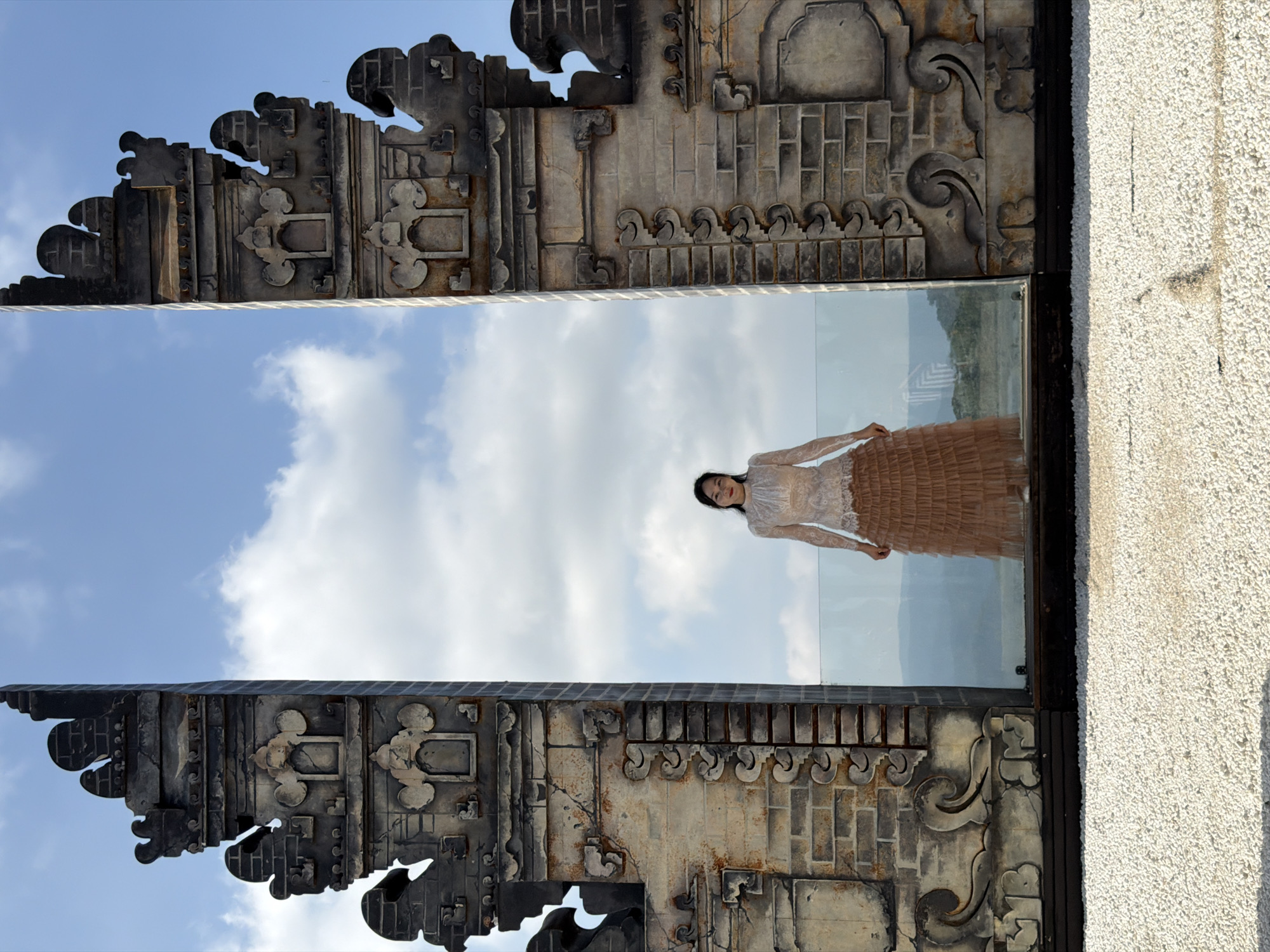 A young Asian woman stands between two ornate, weathered stone gateways, dressed in a white lace top and a flowing, tiered beige skirt. The gateways, constructed with dark brick and decorative grey stone carvings, frame a distant landscape of hills under a bright blue sky with scattered white clouds. The ground in the foreground is covered with small white pebbles. The scene appears to be an outdoor architectural viewpoint, potentially a tourist attraction in Da Lat, Vietnam, known for its French colonial architecture and natural beauty.