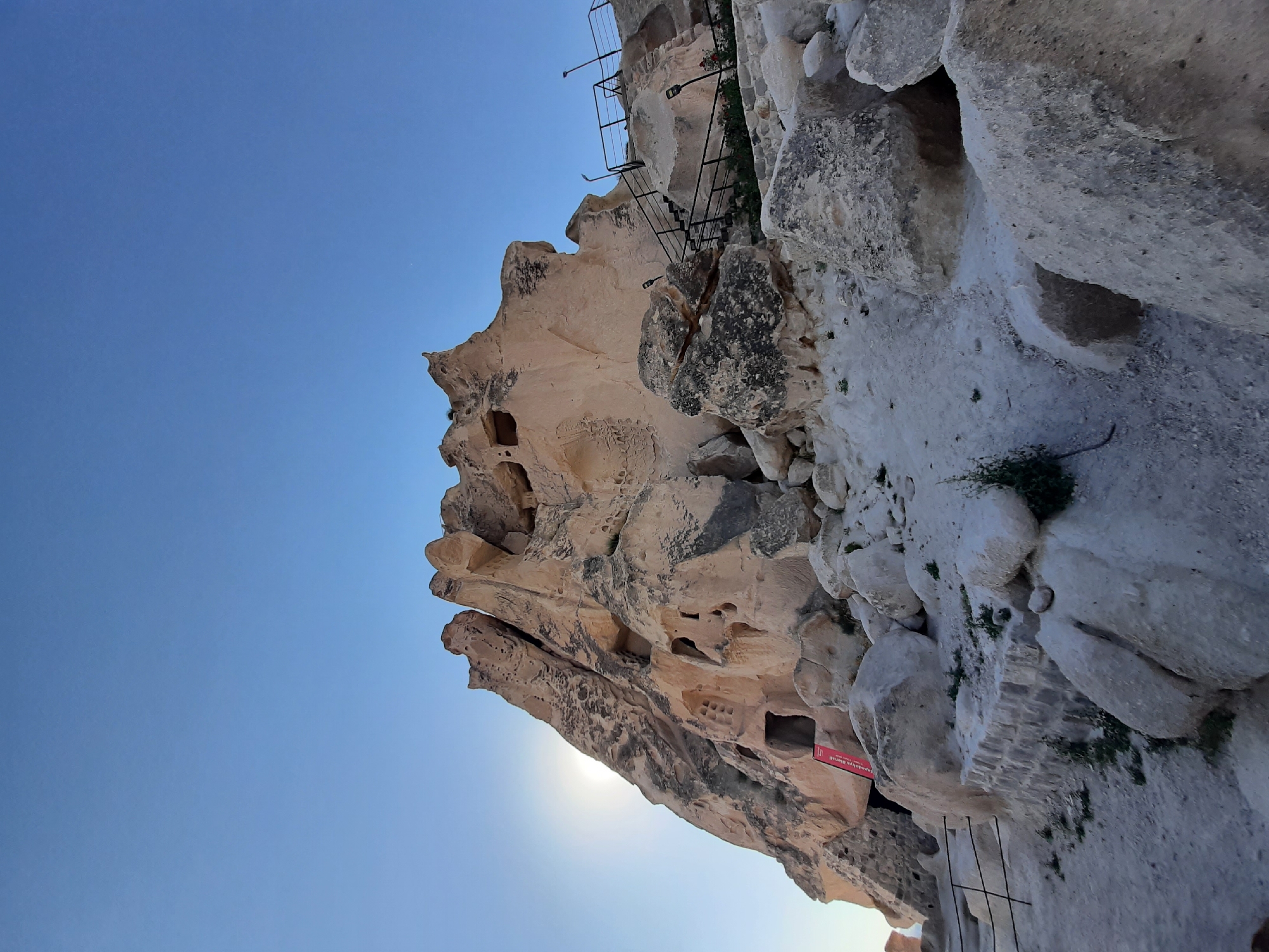The image shows a wide-angle, low-angle view of a rock formation with ancient cave dwellings in Uçhisar Beldesi, Turkey. The sky is clear blue, with the sun partially obscured by a tall, jagged rock spire on the left. The rock formation is light brown and weathered, with numerous openings and carved-out spaces, indicating past human habitation. Several large boulders and smaller stones are scattered at the base of the formation, creating a rocky, uneven terrain.
To the right of the main rock formation, a dark metal staircase with a railing ascends the side of the rock. Several black lampposts are also visible along the staircase and on the path leading up. Small green plants and some red flowers are growing in patches around the base of the stairs and along the path.
In the lower left portion of the image, near the entrance to a cave dwelling, a red and white sign with Turkish text is visible. Black metal railings are also present along the pathways at the lower level. The overall impression is one of ancient history and natural erosion, set against a bright, clear sky. The scene appears to be a tourist site, with the staircase and railings suggesting accessibility for visitors.