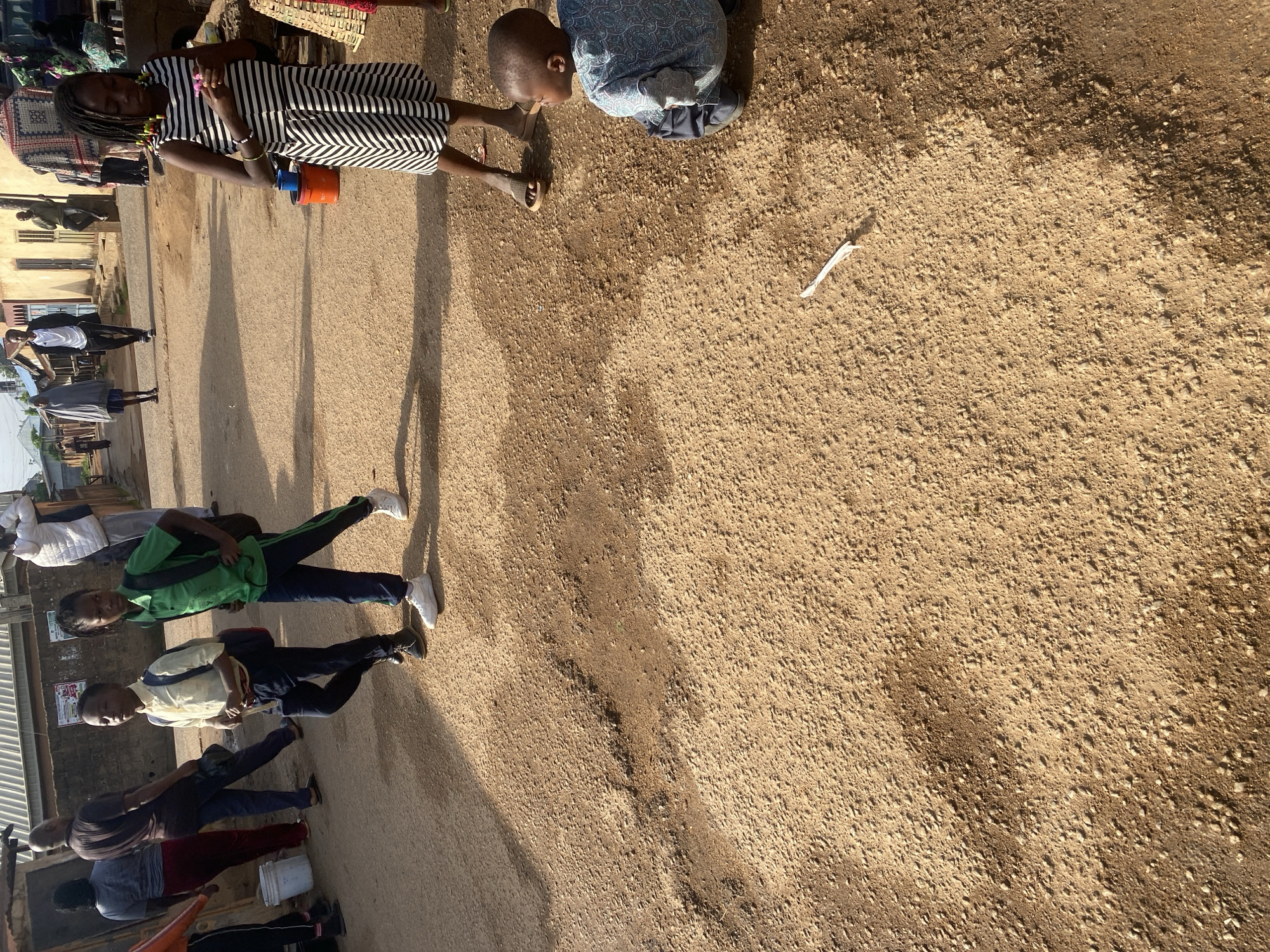 The image captures a street scene in Jos, Nigeria, with a dirt road and a few buildings in the background. In the foreground, a young boy in a blue and white patterned shirt is crouched on the ground, looking to his right. Several other children and adults are visible in the midground and background. Two boys, one in a yellow shirt and backpack and another in a green shirt and backpack, are walking towards the right side of the frame. A girl in a striped dress is standing in the midground, looking down at her hands. Further in the background, a group of people are walking along the street, and some are standing near the buildings. The environment appears to be a somewhat dusty, unpaved urban area.