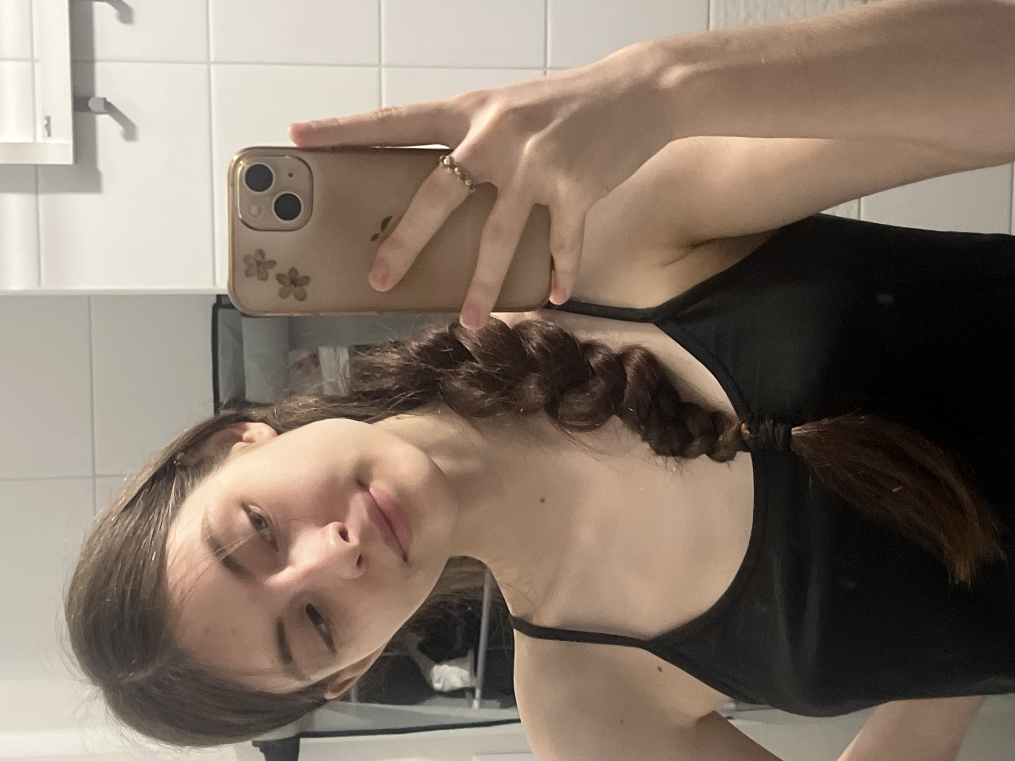A young woman with brown hair braided into a ponytail is taking a selfie in a bathroom. She is wearing a black tank top. The bathroom features white tiled walls and a cabinet with a white door on the wall. The woman is holding a gold iPhone with flower designs on the case. Her hand has a gold ring on one of her fingers. No information is available to determine the location in Saint Petersburg, Russia.
