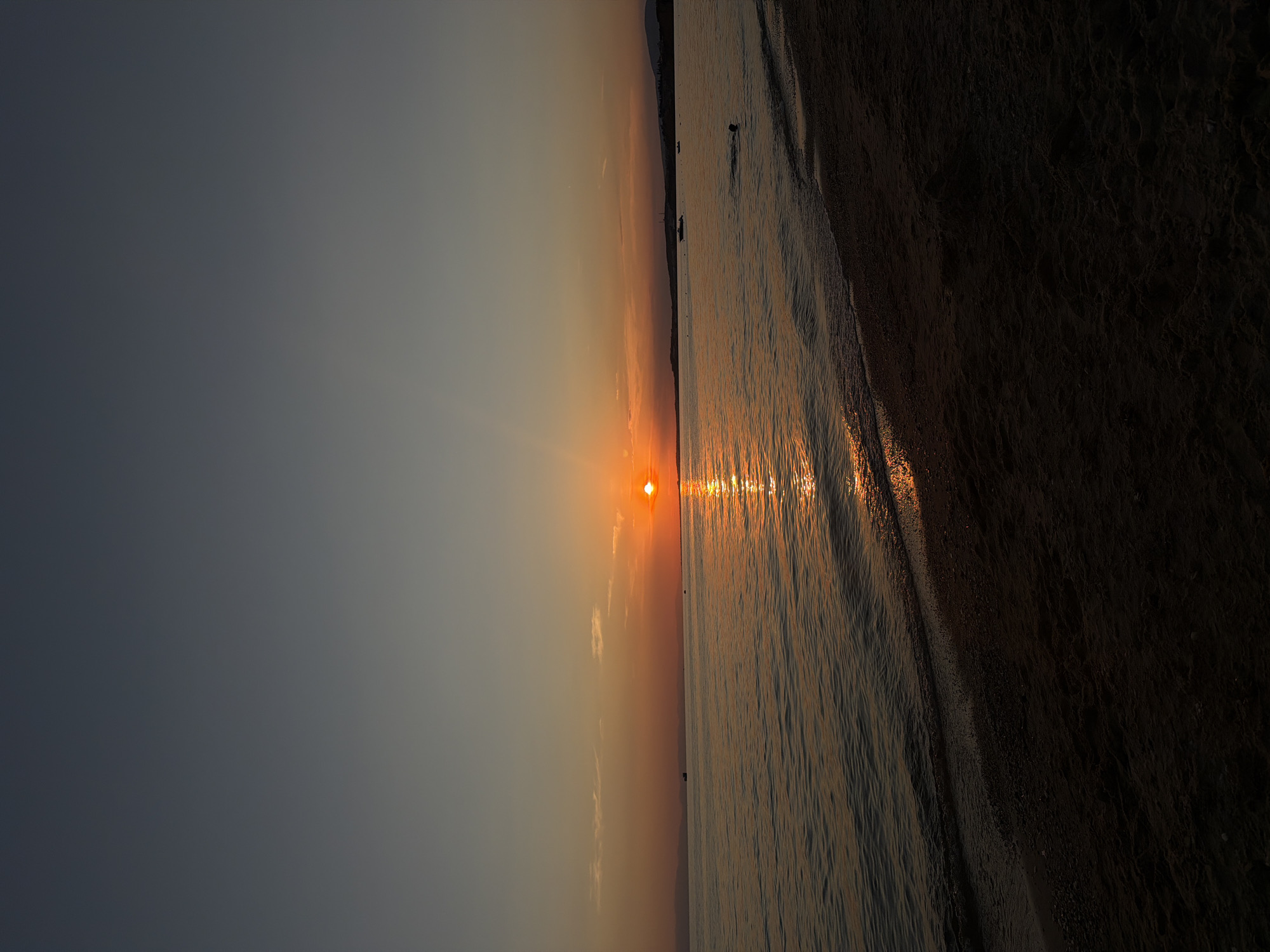 A vertical, full shot captures a tranquil sunset over the calm waters of the Aegean Sea at Πόρτες, Greece. The sun, a radiant orange orb, is positioned slightly left of center on the horizon, casting a shimmering golden reflection across the water's surface. The sky transitions from a deep, dusky blue at the top to a warm gradient of orange and yellow near the horizon. A sliver of land, with faint mountain silhouettes, forms the distant shoreline.
In the foreground, a sandy beach occupies the bottom third of the image, dark and textured with small pebbles. The gentle waves lap at the shore, creating a subtle line where the water meets the sand.
Further out in the sea, a lone swimmer is visible, their head and shoulders breaking the surface. A few small boats are dotted in the distance, adding to the sense of serenity. The overall atmosphere is peaceful and serene, bathed in the warm glow of the setting sun.