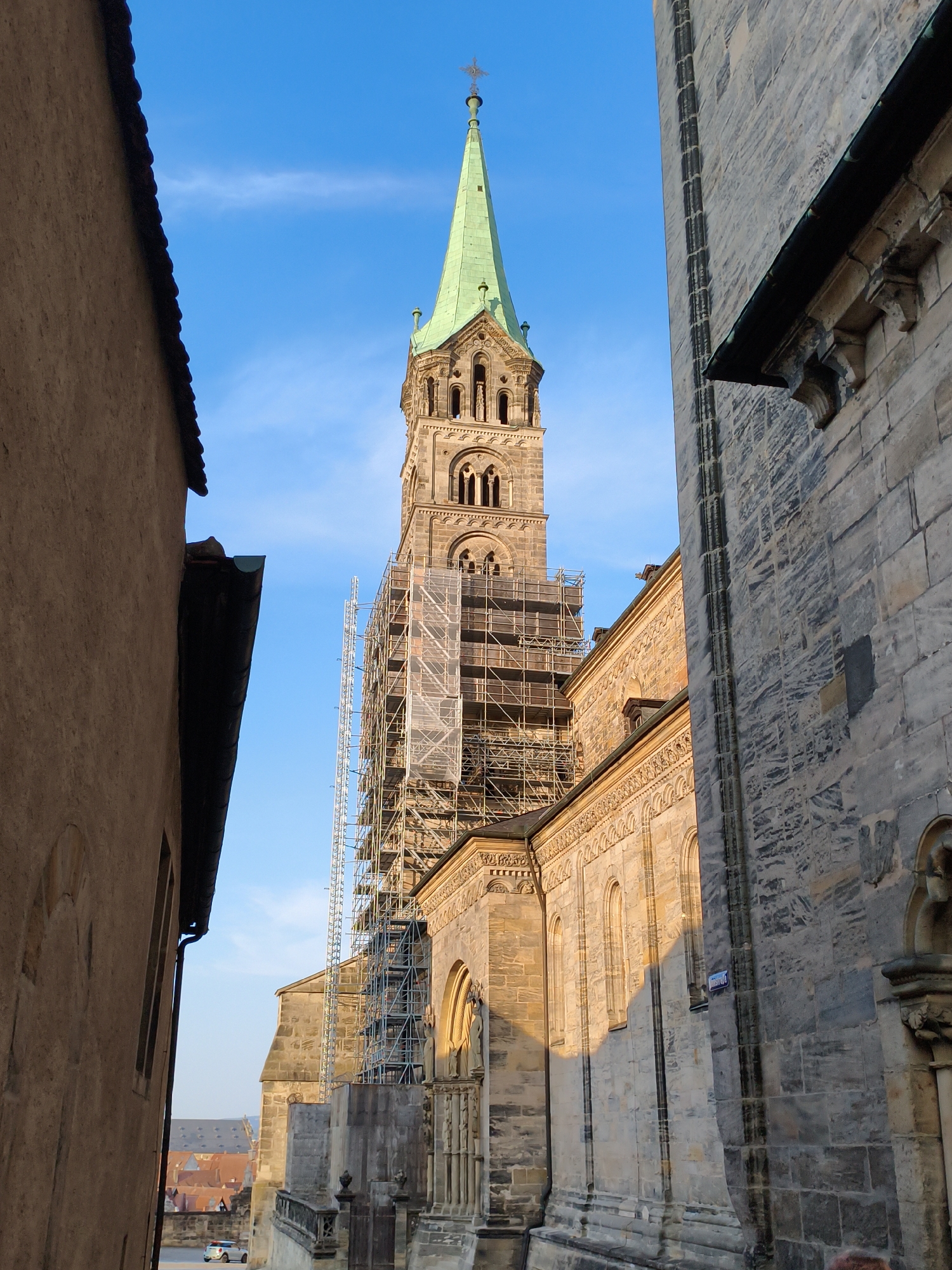 This image captures a view of the Bamberg Cathedral (Bamberger Dom) in Bamberg, Germany, with scaffolding on its tower. The perspective is from a narrow alleyway between two buildings, looking towards the cathedral's spire, which is topped with a cross. The cathedral's stone facade is visible, showcasing its architectural details and arched windows. The tower is partially covered by scaffolding, suggesting ongoing restoration or maintenance work. Sunlight illuminates the right side of the cathedral. The sky is blue with a few wispy clouds. In the background, partially visible through the gap in the buildings, are rooftops of other structures in the city. The left side of the image is dominated by the textured, light brown wall of an adjacent building, with a dark roofline visible along its top edge.