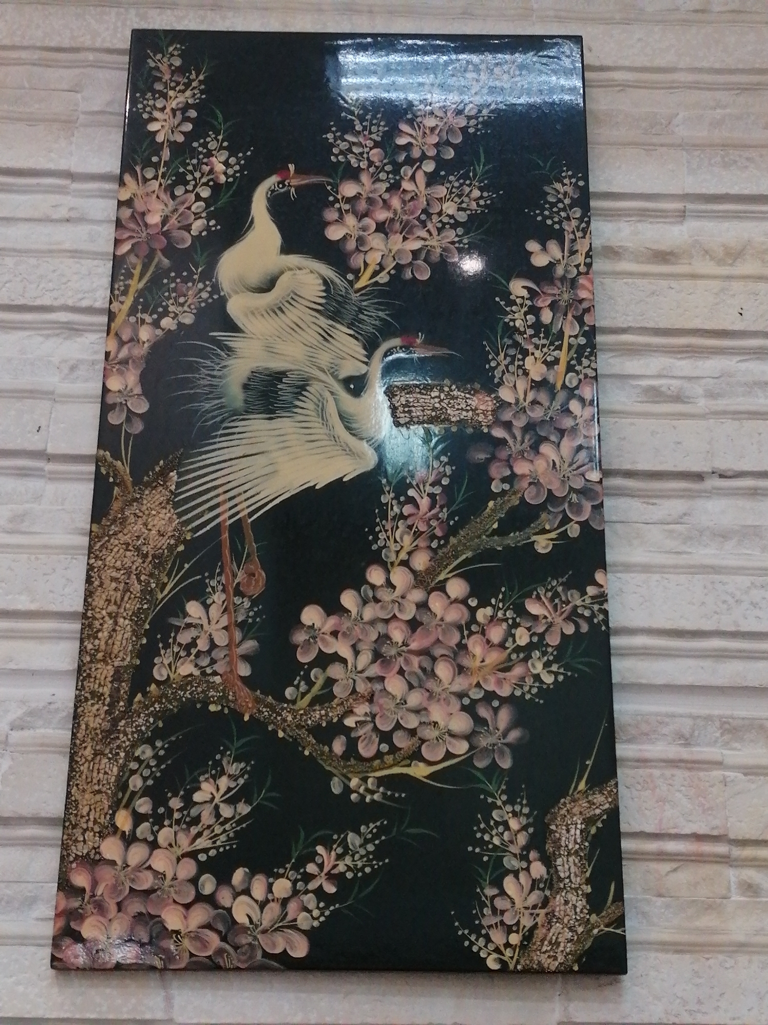 The image shows a decorative panel featuring two cranes amidst cherry blossoms. The panel is dark with intricate detailing of white and pink flowers and two elegant cranes. The cranes are depicted in white with red accents on their heads. The background is black, providing a stark contrast to the colorful elements. The panel is leaning against a textured, light-colored wall, which appears to be made of bricks or a similar material with horizontal lines. The overall impression is of an ornamental piece with a traditional artistic style. This panel is likely displayed indoors, given its presentation against a wall. There is no indication of the specific location within Ho Chi Minh City, Vietnam.