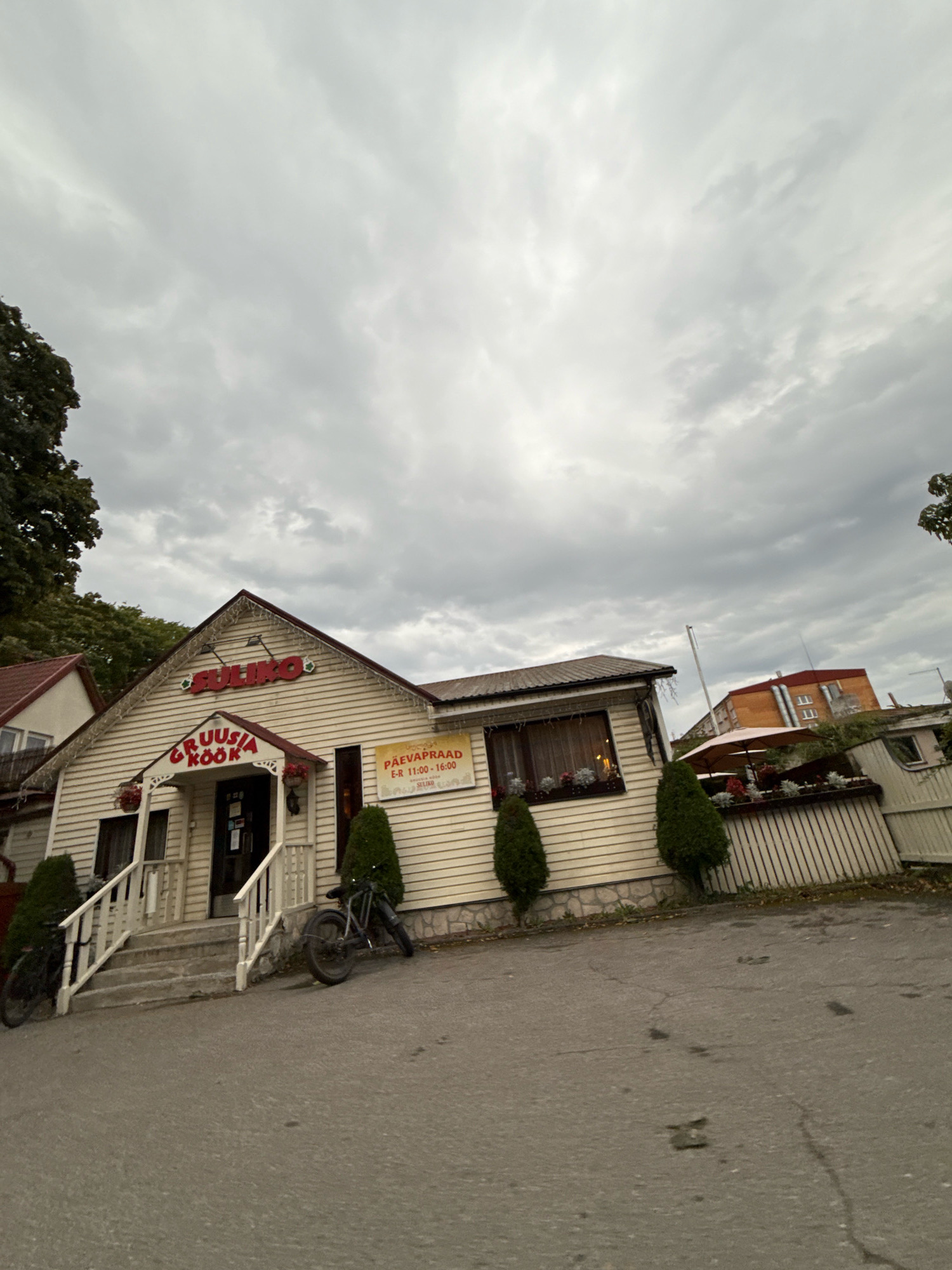 The image depicts the exterior of a restaurant named "SULIKO" with the signage "GRUUSIA KÖÖK" above the entrance. A sign indicating "PÄEVAPRAAD E-R 11:00 - 16:00" is visible on the right side of the building. The building is light-colored with a red roof. There are two bicycles parked outside the entrance, one on the left and one on the right. The scene is set on a paved area in front of the restaurant, with some trees and other buildings in the background. The sky is overcast. The restaurant is located in Tallinn, Estonia.