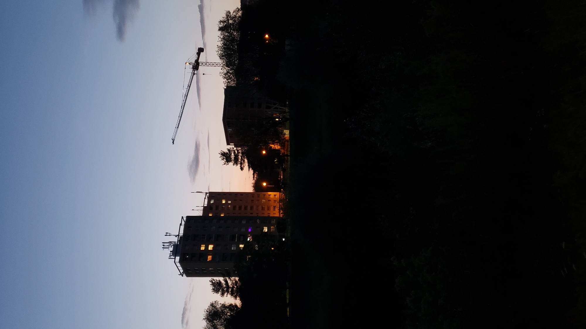 This image depicts a twilight scene in Prešov, Slovakia, with a focus on residential buildings and a construction crane. In the background, two tall apartment buildings are visible, with some windows illuminated, suggesting occupancy. To the right of the apartment buildings, a construction crane stands silhouetted against the darkening sky, indicating ongoing development or construction work. Trees and foliage frame the scene in the foreground and midground, adding a natural element to the urban landscape. The sky transitions from a pale blue near the horizon to a deeper twilight hue above, with a few wispy clouds present. The overall atmosphere is calm and serene, characteristic of the transition from day to night.