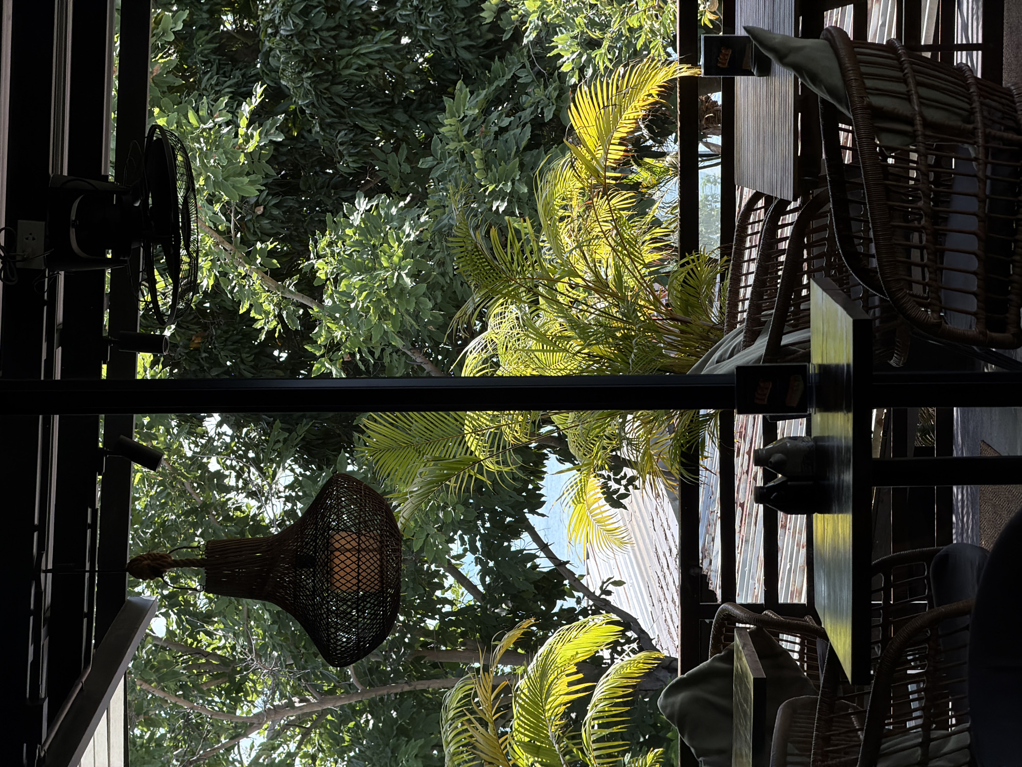 The image depicts an outdoor seating area at what appears to be a restaurant or cafe, possibly located in ตำบลเกาะพะงัน, Thailand. The scene is viewed from a covered section, looking out towards lush green foliage, with glimpses of the ocean in the distance. In the foreground, several tables and chairs are visible. The tables are dark wood, and the chairs are made of woven rattan with light-colored cushions. On one table, two dark bottles, likely for condiments, are placed. A large, woven pendant light fixture hangs from the ceiling of the covered area, and a ceiling fan is also mounted above. The background reveals a slightly weathered, corrugated metal roof structure, beyond which is a dense canopy of green trees and the blue expanse of the sea. The lighting suggests it is daytime, with sunlight filtering through the leaves and casting shadows. There are no people visible in the image.