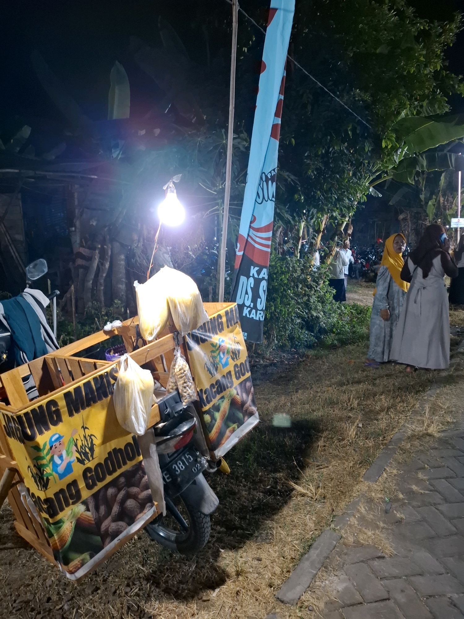 In Ringin Anom, Indonesia, at night, a food cart is displayed. The cart is wooden and yellow, with the words "UNUNG MAKAN" and "KACANG GODHO" printed on its sides. The cart is adorned with bags of corn and peanuts. A motorcycle is attached to the cart, with a license plate that reads "E 3815 02". The background shows trees and a street with parked motorcycles. Two women, dressed in traditional clothing, are seen standing near the cart, observing the surroundings. The scene is illuminated by a single light bulb hanging from a pole.