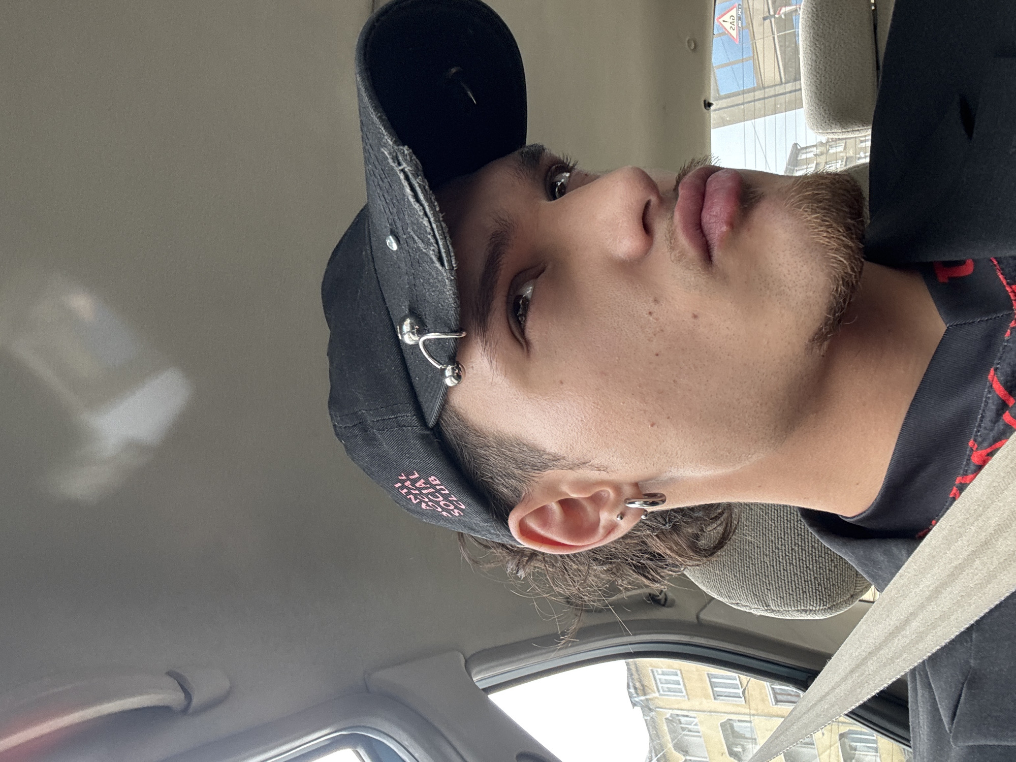 A young male is depicted from the mid-chest up, seated in the passenger side of a vehicle, angled slightly to his left and looking upwards. He has dark, curly hair visible beneath a black, distressed baseball cap with a frayed brim. The cap is embroidered with pink "ANTI SOCIAL CLUB" text and adorned with several silver-toned metallic piercings and studs. His visible right ear features a small silver stud and a larger silver hoop earring. He has prominent eyebrows, dark eyes, light stubble/scruff on his cheeks, a thin mustache, and a reddish-brown goatee. A small blemish is present on his lower lip. He is wearing a black jacket or blazer over a dark shirt with a red pattern. A beige seatbelt is secured across his chest. The vehicle's interior features a light-colored headliner and grey textured fabric on the headrest behind him.
Through the windows, an urban environment is visible, characteristic of an older residential area in Sofia, Bulgaria. A multi-story residential building with a faded yellow facade and numerous rectangular windows, some with white air conditioning units, is prominent in the background. Through the rear window, a red triangular road sign with a white background and black symbol (likely a general danger warning) is visible, along with Cyrillic text, including "ГАЗ" (GAS), on what appears to be another sign or a reflection, confirming the Bulgarian context. The sky is overcast.