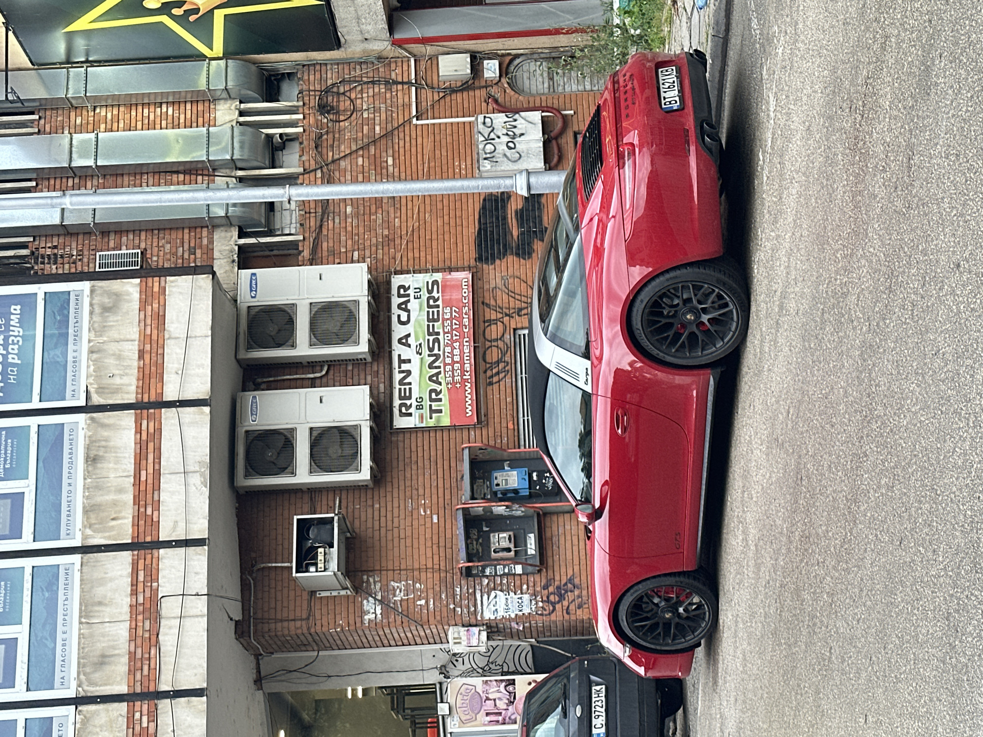 A red 2016 Porsche 911 Targa 4 GTS is parked on a street in Veliko Tarnovo, Bulgaria, alongside a black car and two vintage payphones. The car is a vibrant cherry red with a silver roof and black wheels. The rear of the vehicle displays a Bulgarian license plate that reads "BT 1621 KB". The background reveals a brick building with several air conditioning units mounted on its facade. Graffiti adorns the brickwork. A "Rent a Car" sign is visible above the payphones. The street appears to be paved and there is some overgrown vegetation on the right side of the image. The scene suggests an urban setting with a mix of old and new elements, typical of a European city.