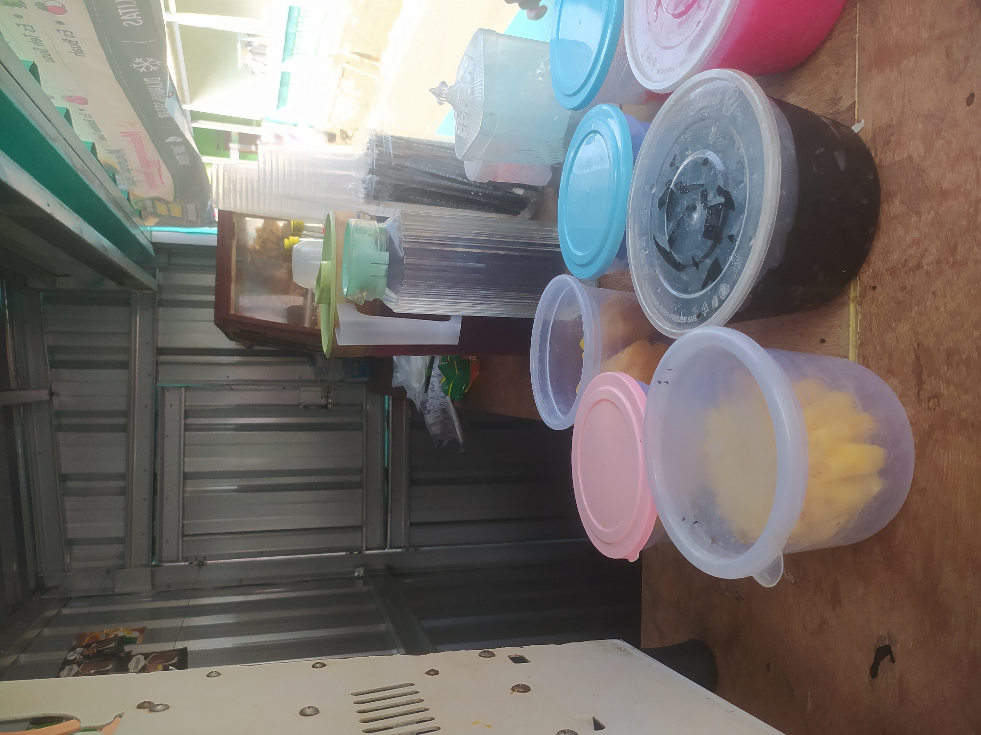This image depicts a food stall in Lopait, Indonesia. The stall appears to be outdoors, with a corrugated metal structure forming the back wall and part of the roof. On a wooden counter, various containers are arranged, suggesting prepared food or drinks.
In the foreground, several plastic containers are visible. One container on the left has a pink lid and appears to hold a yellow, shredded ingredient, possibly fruit. Next to it is a larger, translucent plastic container with a light purple rim, containing a similar yellow shredded material. To the right, a clear plastic container with a blue lid holds a portion of brown liquid or semi-solid. Another blue-lidded container is partially visible. Further to the right, a container with a pink lid contains a pink substance.
Behind these containers, several larger plastic jugs and containers are present. One tall, clear plastic jug with a light green lid contains a dark liquid. Another tall, clear plastic container to its right holds dark, elongated items, possibly straws or dried ingredients. A square, clear plastic container with a decorative lid, resembling a sugar dispenser, is also on the counter. Several disposable plastic cups are stacked behind these items.
The background shows the interior of the stall, including corrugated metal walls and framing. A banner or sign with text, partially obscured, is visible at the top of the stall. Some food packaging or wrappers are attached to the back wall. The overall impression is of a functional, albeit somewhat cluttered, street food stall. No people are visible in the frame.
