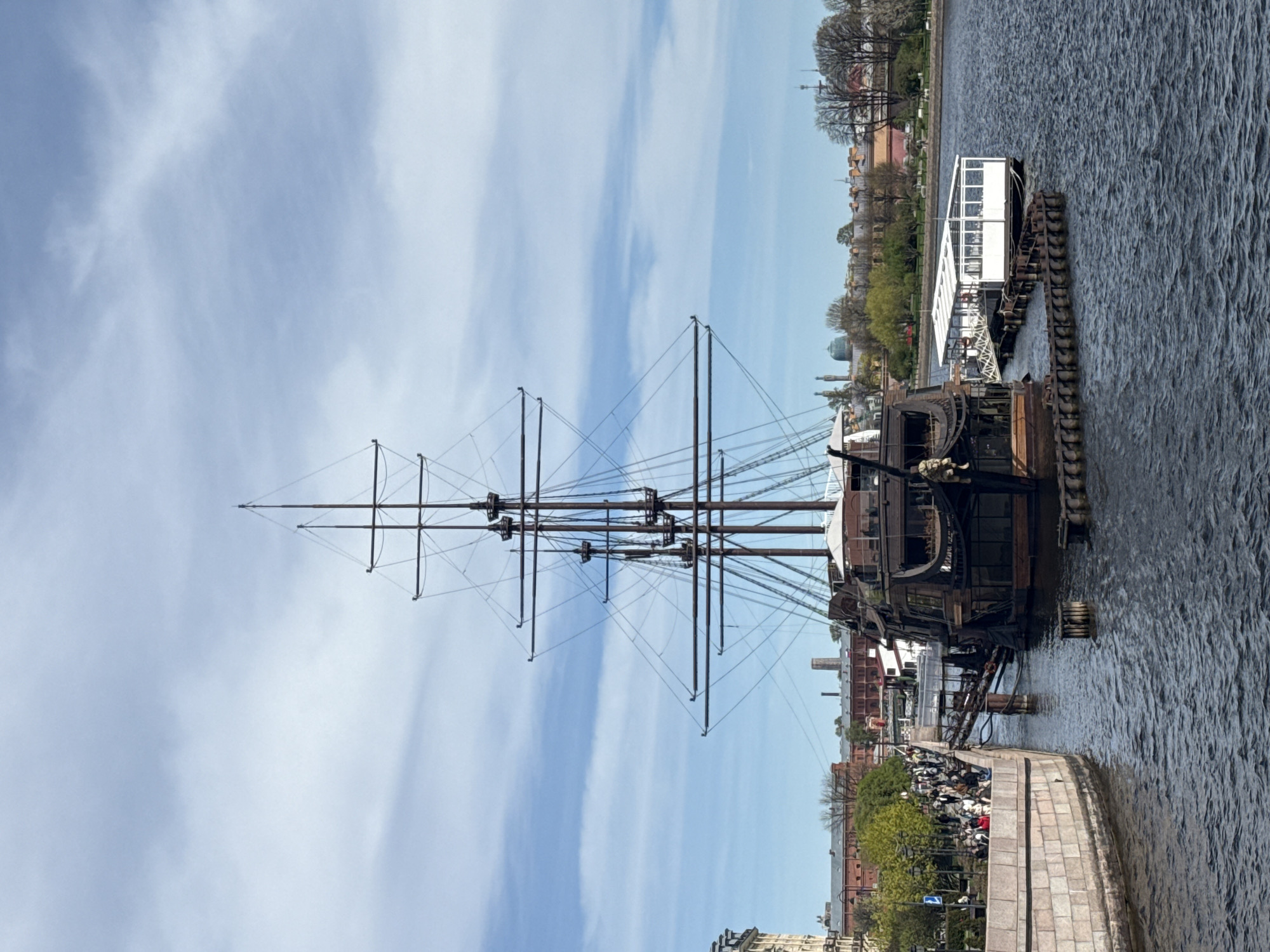 The image shows a tall ship, likely a replica or a historical ship, docked on a waterway. The ship's mast dominates the composition, with rigging creating a network of lines against the blue sky with streaky clouds. The hull of the ship is dark and appears weathered.
People are gathered along the embankment, viewing the ship. A modern white boat is docked nearby. The waterway is dark with ripples.
The location is most likely Saint Petersburg, Russia, as the city is known for its waterways and historical maritime connections. The architectural style of the buildings along the embankment is consistent with Saint Petersburg's historical architecture.
