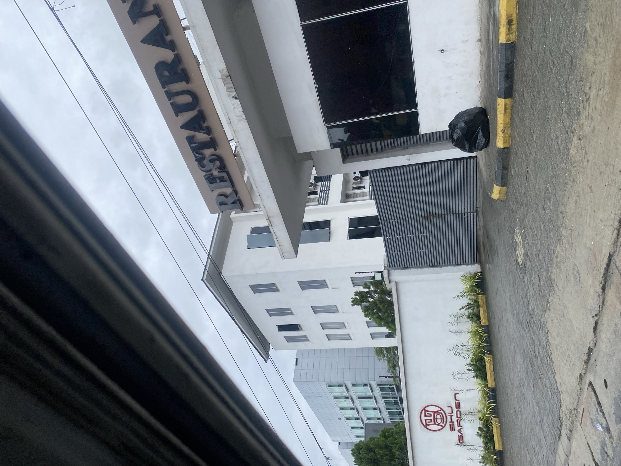 The image shows the exterior of a restaurant on Victoria Island, Nigeria. A sign above the entrance reads "RESTAURANT" in large, dark, uppercase letters. Below the sign, a building with a white facade features large, dark-tinted windows. To the left of the restaurant entrance, a white wall displays the logo and name "SHU GARDEN". Adjacent to the wall is a grey, corrugated metal gate. The ground is paved with interlocking bricks, and a black trash bag sits on the curb. In the background, two modern multi-story buildings are visible, one white and one silver, with numerous windows. The sky is overcast.