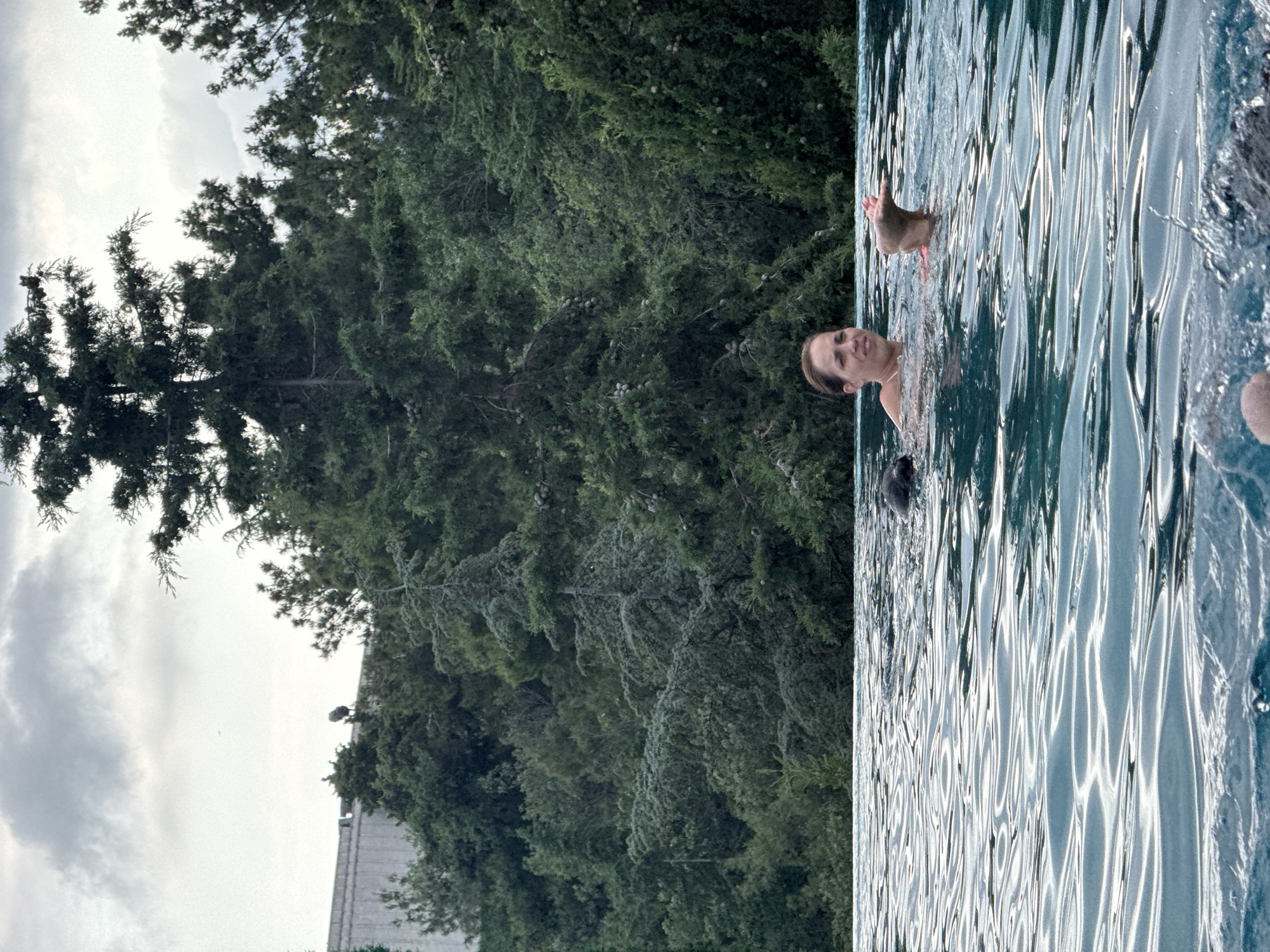 A woman is swimming in a pool. The woman is in the center of the frame, with her head and shoulders visible above the water. She is looking to the left and smiling. Her left arm is extended, and her hand is out of the water. Behind her, in the water, is a black dog, partially submerged. The pool is surrounded by lush green trees, and a building is visible in the background. The sky is overcast. The scene appears to be taking place in a private residence or a resort. The location is identified as Yalta, Ukraine.