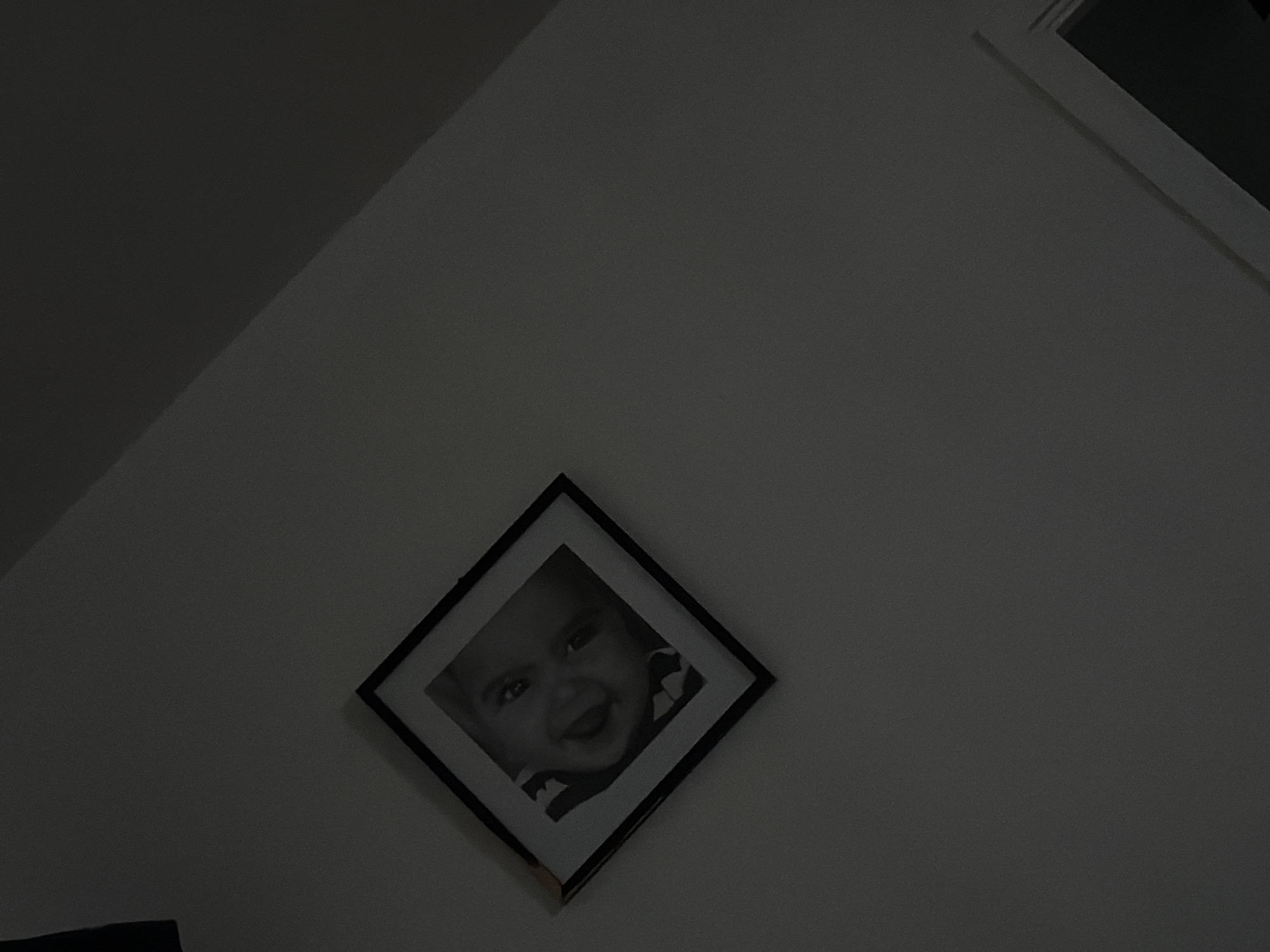 A black and white framed photograph of a baby is hanging on a wall in a dimly lit room. The photograph is square and tilted so it appears like a diamond. The baby in the photograph is smiling and looking directly at the camera. The background of the photograph is dark. The wall the photograph is hanging on is white. The image is taken from a low angle, looking up at the photograph. There is a shadow cast on the wall to the right of the photograph. The room appears to be a bedroom or living room.