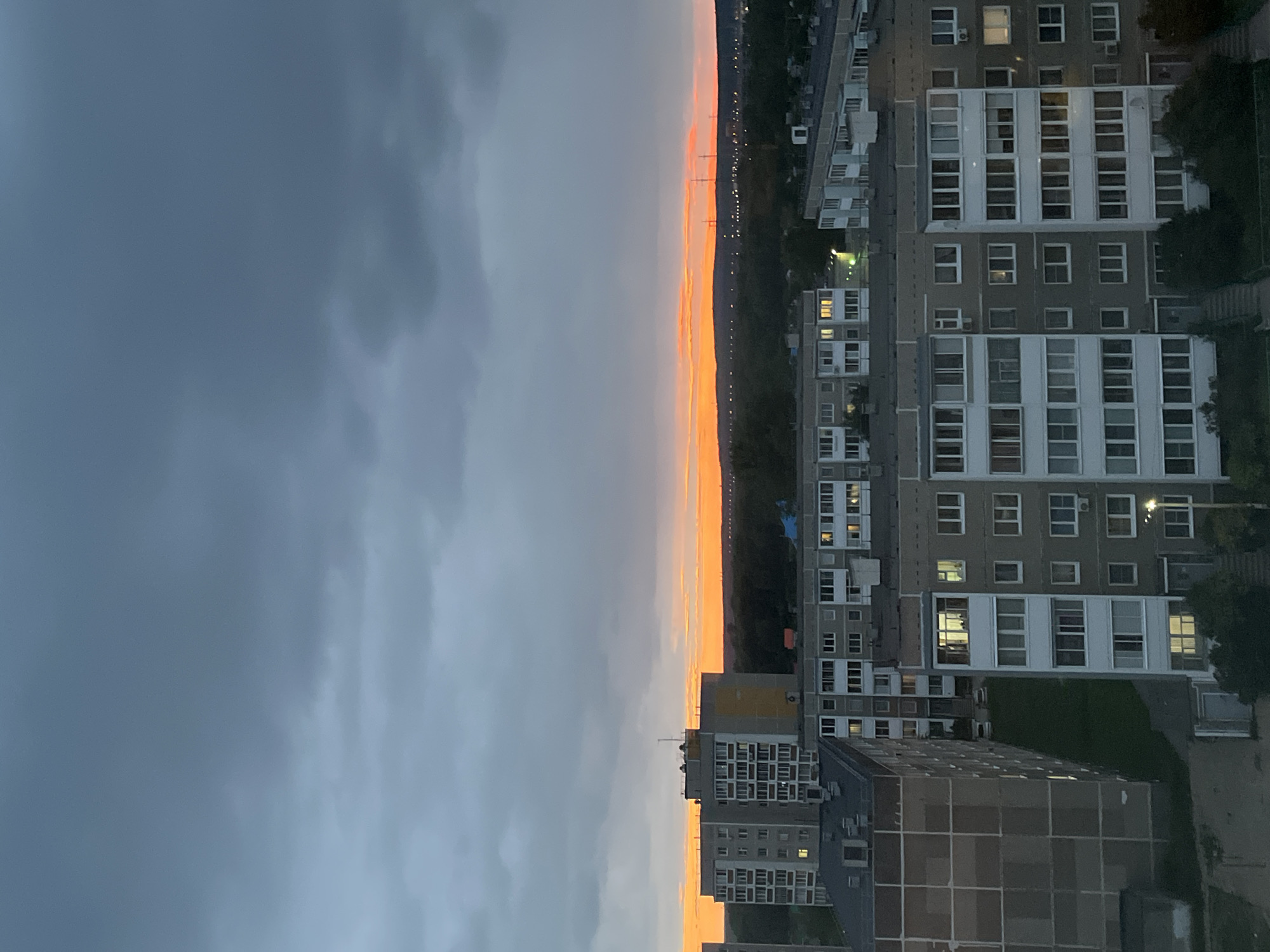 An aerial view captures apartment buildings against a dramatic sky at dusk in an urban setting. The sky is dominated by dark, cloudy formations in the upper half, transitioning to a vibrant band of orange and yellow light along the horizon where it meets a dark, tree-lined landscape. Below the sky, numerous multi-story apartment buildings with many windows are visible. Some windows are illuminated, indicating indoor activity. In the foreground, closer apartment buildings are detailed, with visible facades and architectural features. The environment appears to be a residential area within a city.