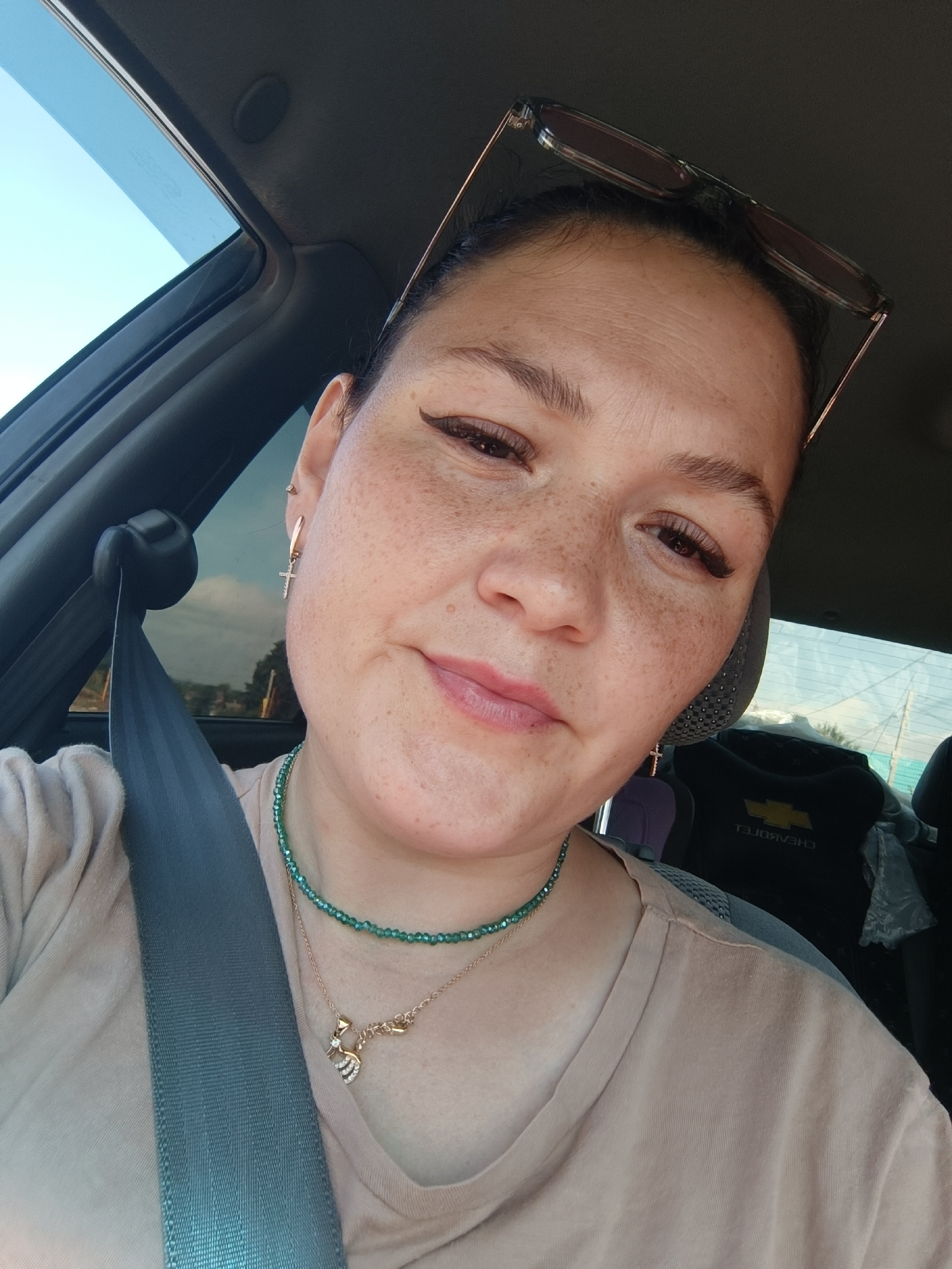 A woman is captured in a close-up, selfie-style shot from inside a vehicle, possibly a car. She is wearing a light-colored t-shirt and is adorned with two necklaces, one green beaded and another with a golden pendant. A cross-shaped earring dangles from her right ear. Her gaze is directed towards the camera, with a slight smile on her lips. Sunglasses are perched on top of her head. The background reveals the interior of the car, including a seatbelt, a headrest, and a portion of a black item with a gold Chevrolet logo. The window of the car shows a blurred glimpse of the exterior, suggesting movement. No specific landmarks or elements identifying the exact location within Chaikovsky, Russia, are discernible from the image.