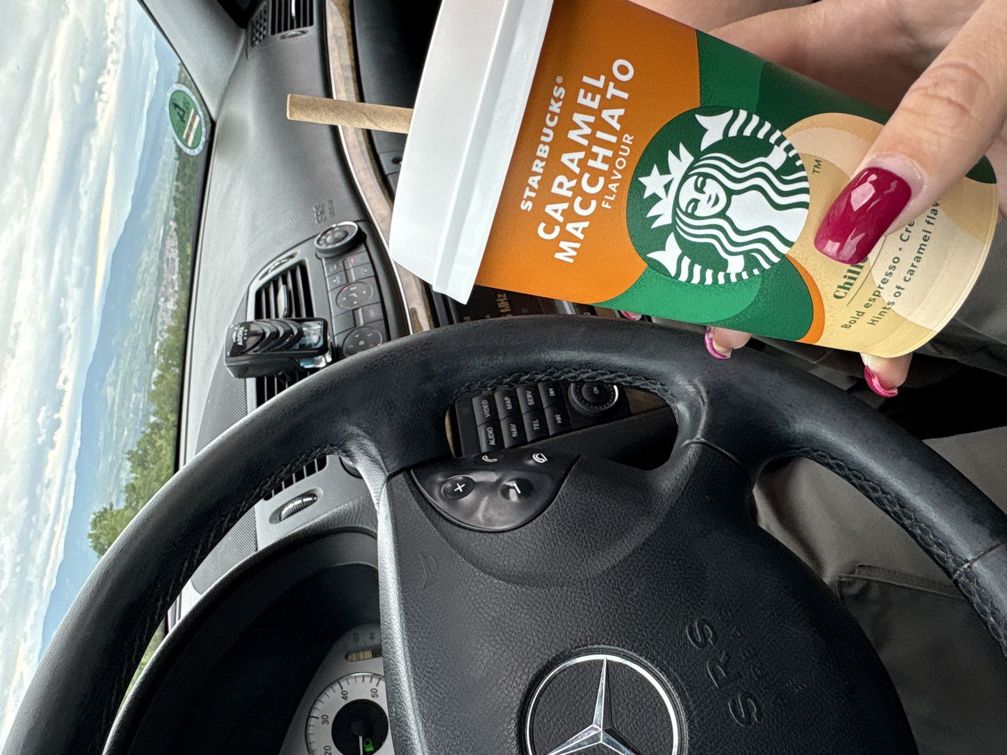The image, taken from within a Mercedes-Benz vehicle, shows a person holding a Starbucks Caramel Macchiato cup. The interior of the car includes the steering wheel, dashboard, and a partial view through the windshield. The steering wheel is black, with the Mercedes-Benz logo prominently displayed in the center. The dashboard features various controls and an integrated navigation system. Through the windshield, a scenic landscape is visible, including mountains and a clear sky. The Starbucks cup is orange and green, with the company logo and the words "Caramel Macchiato Flavour." The person's hand, with a painted nail, is holding the cup. The landscape visible through the car windshield appears to be the area around Marulevo, Bulgaria.