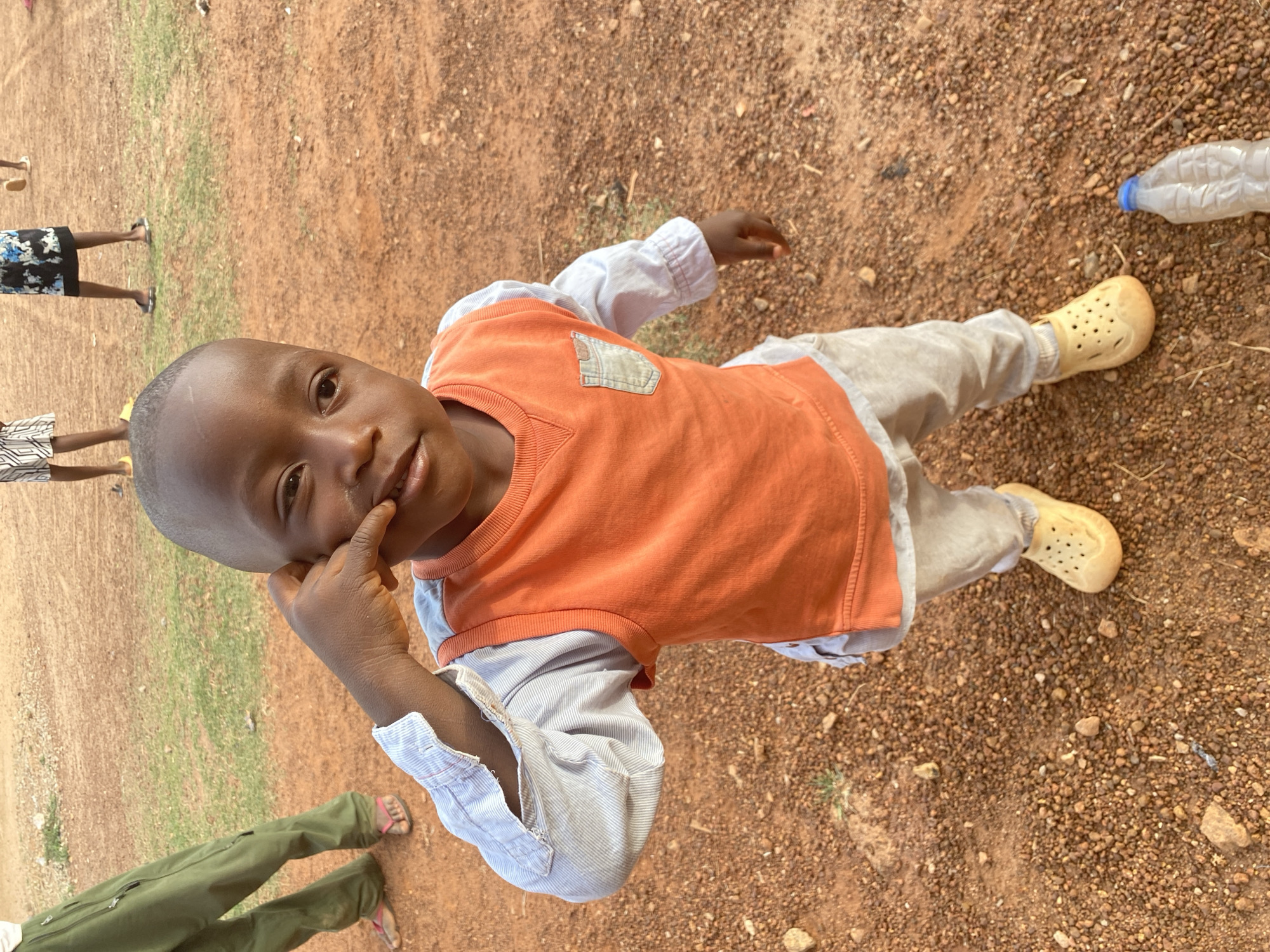 An overhead shot captures a young child standing outdoors on reddish-brown gravel and dirt, with sparse patches of green grass visible. The child, with dark skin and a shaved head, looks directly at the camera with a gentle expression, one finger resting near their cheek. They are dressed in an orange sleeveless top with a denim-look pocket over a light blue and white striped long-sleeve shirt, light-colored pants, and yellow Crocs-style shoes.
In the background, the lower halves of at least three other individuals are visible, also standing on the same ground. One person wears black and white patterned shorts and dark flip-flops, another has blue and white patterned shorts and dark flip-flops, and a third wears green cargo-style pants and pink flip-flops. An empty, clear plastic water bottle with a blue cap lies on the ground near the child's feet. The scene appears to be brightly lit, suggesting daytime. This outdoor gathering takes place in Bukuru, Nigeria. No visible text is present in the image.