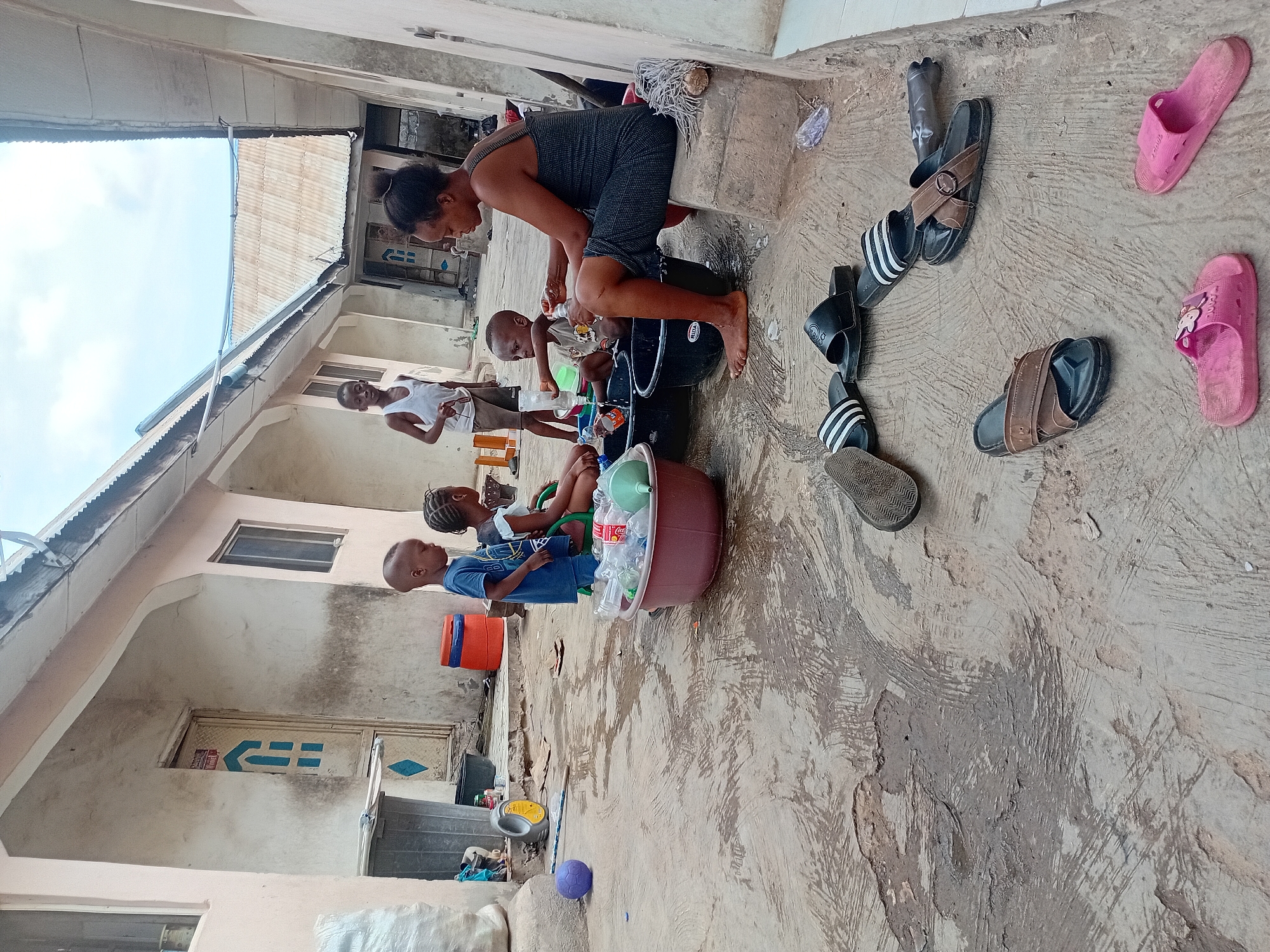 The image depicts an outdoor scene in what appears to be a residential courtyard or alleyway in Lokoja, Nigeria, during the daytime, likely under an overcast sky. Several individuals, including an adult woman and multiple children of varying ages, are gathered, engaged in what appears to be communal washing or bathing.
In the foreground, an adult woman, dressed in a dark top and shorts, kneels next to a black bucket, attending to a small child seated within it. Another child stands nearby, holding a plastic bottle, while other children are visible further back, some interacting with a large brown basin filled with plastic bottles. The ground is a textured concrete surface, showing signs of dampness and wear.
Various objects are scattered on the ground, including several pairs of sandals and slippers: black and white striped "ADIDAS" brand slides, brown leather-style slides, and pink "PINK" brand slides. Other items include plastic bottles (one clearly identifiable as "Coca Cola"), a purple ball, and a white mop head. One of the black buckets has "RITA" printed on it.
The background features multi-story residential buildings with light-colored rendered walls and windows. Corrugated metal roofing is visible on adjacent structures against the sky. A blue door or gate in the mid-ground displays a distinctive blue and white geometric symbol. The scene conveys a sense of everyday domestic activity in a communal outdoor space.