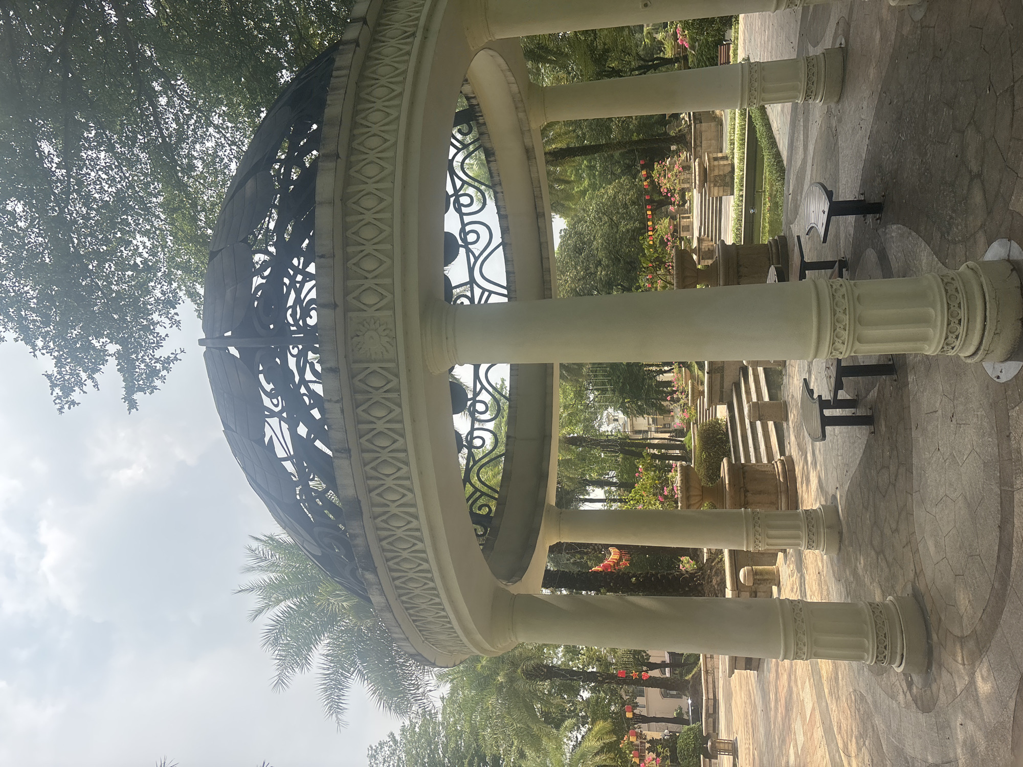 A domed gazebo with white, fluted columns stands in a paved courtyard surrounded by greenery. The gazebo's roof is made of dark metal with intricate scrollwork. In the background, palm trees and other lush vegetation are visible. Red and yellow lanterns hang from some of the trees, suggesting a festive atmosphere. Benches and tables are placed within and around the gazebo. A body of water with plants and a small bridge can be seen in the distance, along with stone steps leading up to a raised area with planters. The scene appears to be a public park in the Nanhai District, China.