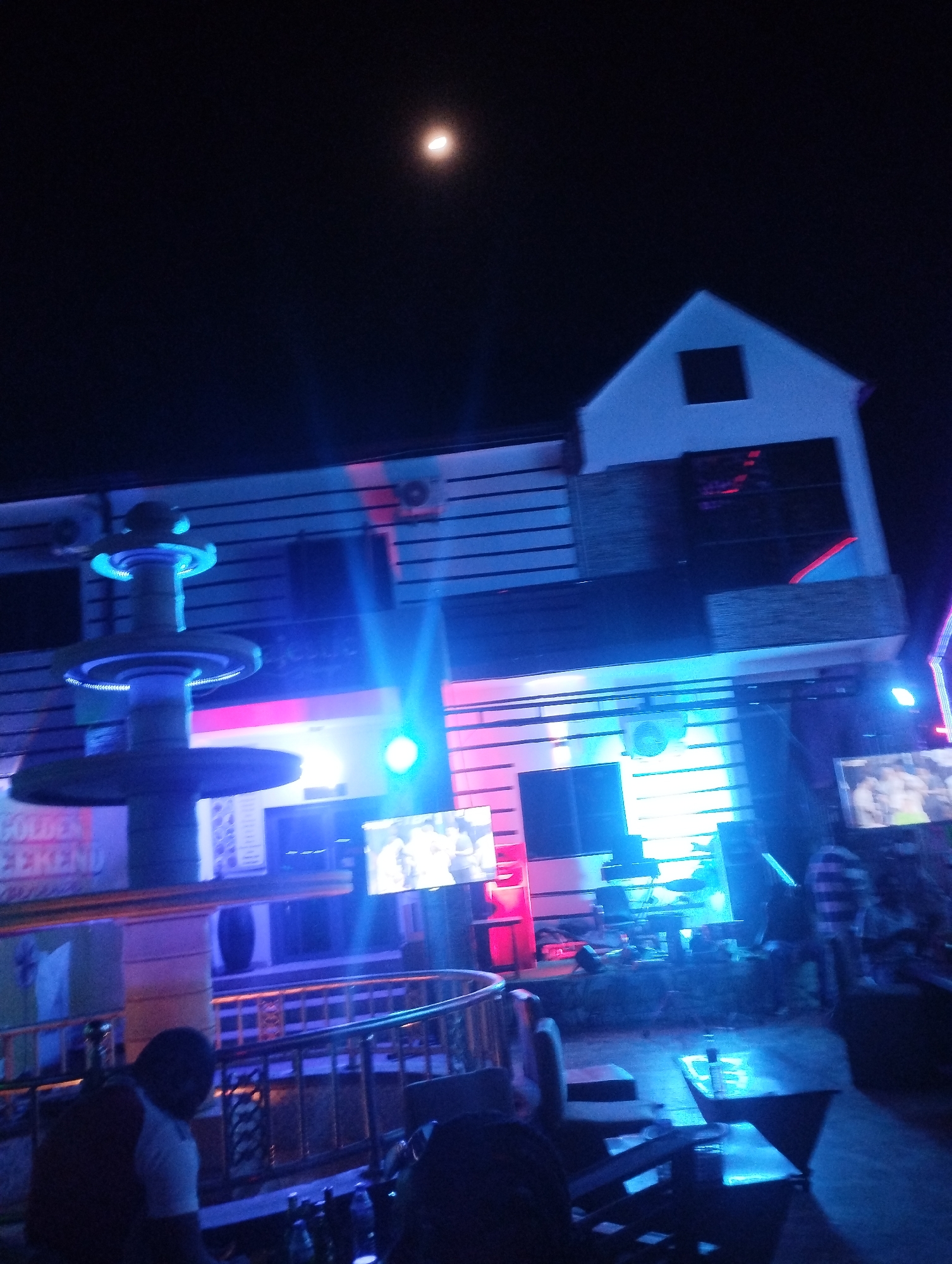 At night in Tumfure, Nigeria, a two-story building is illuminated with blue and red lights. The building has a pointed roof with a square window. Below, the building has multiple windows and at least two air conditioning units are mounted on the exterior. There are at least two televisions showing video content. A tiered fountain is located in front of the building. People are present near tables and chairs, suggesting an outdoor seating area. The moon is visible in the dark sky.
