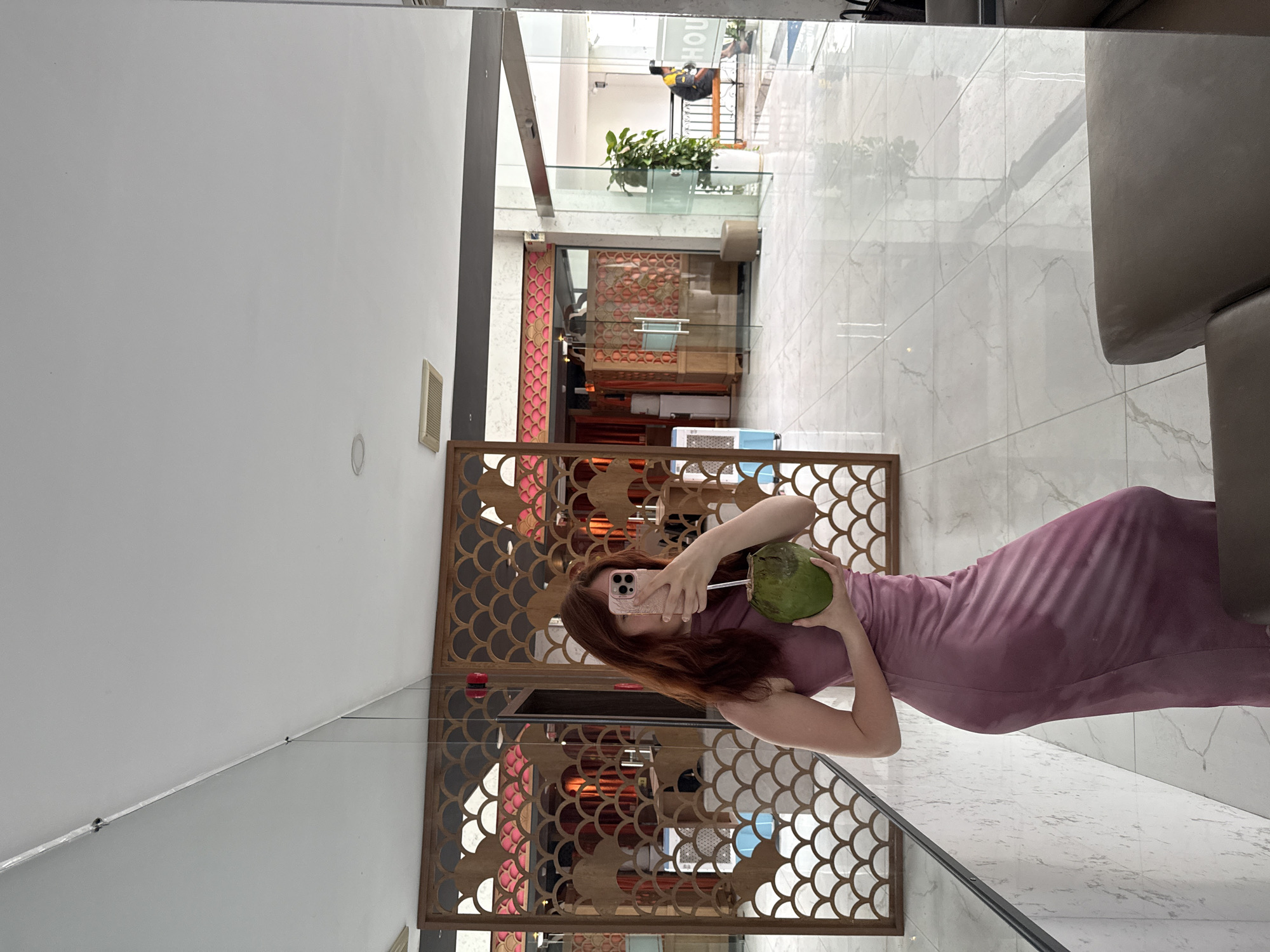 A woman in a pink dress is taking a photo of herself in a mirror, holding a coconut. The mirror is next to a decorative wooden screen. The location appears to be a hotel lobby or similar establishment in Bac Nha Trang Ward, Vietnam, evidenced by the interior design and architectural elements visible in the background. The lobby has a light color scheme, a decorative wall with red and gold accents, and clear tiled flooring.