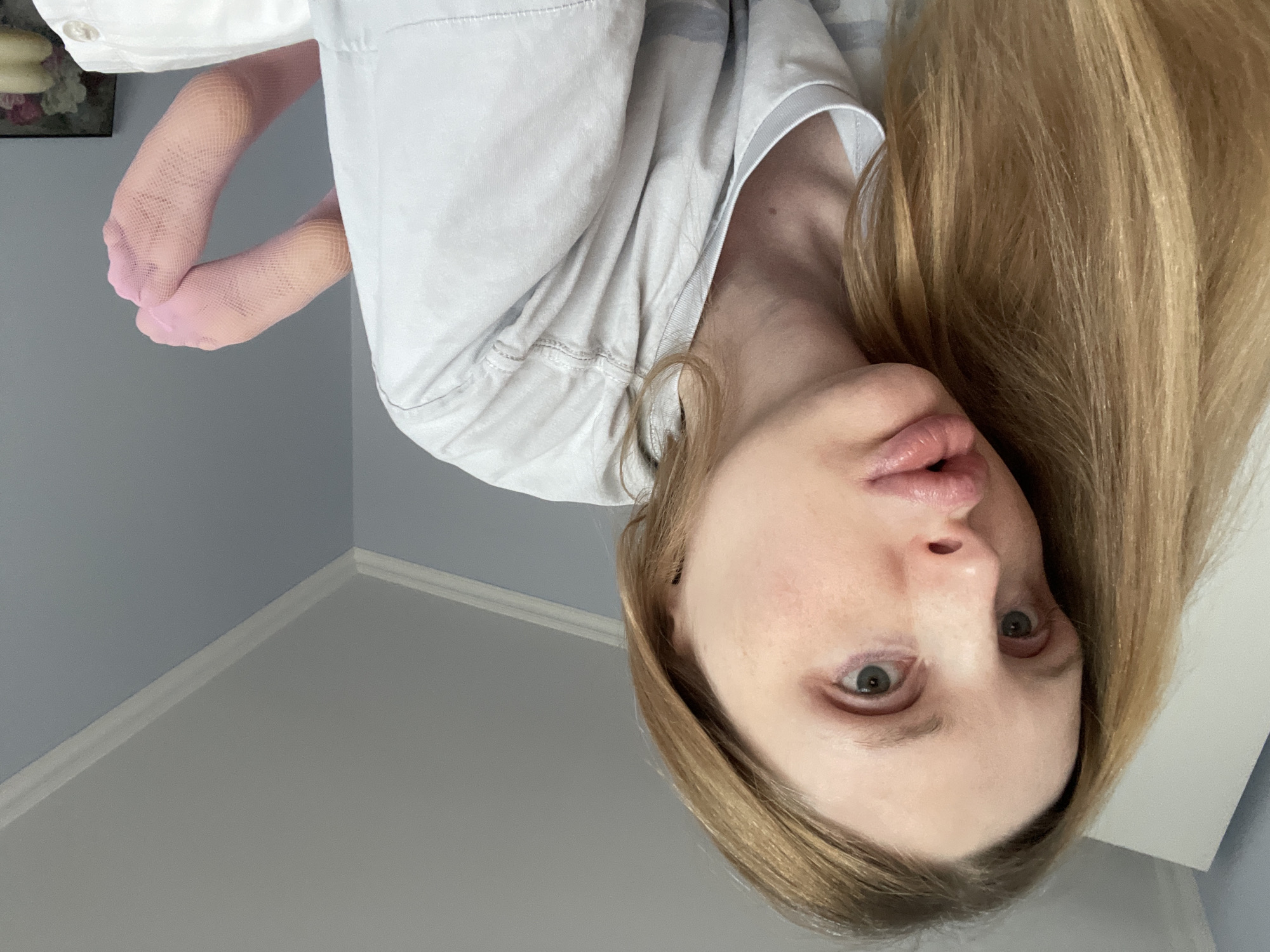 A close-up, low-angle selfie shows a person with long blonde hair lying on their side on a bed. The person is wearing a light grey t-shirt and pink fishnet stockings. Their face is turned to the left, with their lips slightly pursed. The background shows a light blue wall with white crown molding and a white closet door. The scene appears to be indoors, likely in a bedroom. No specific location in Yablonovskiy, Russia, is identifiable from the image.