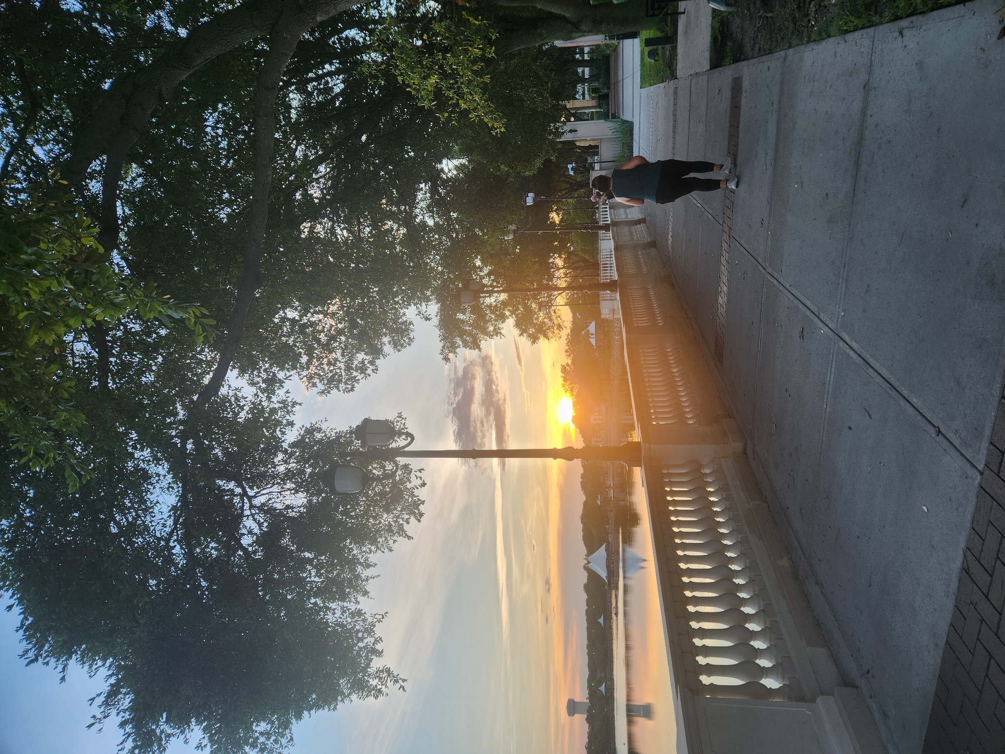 A person jogs on a paved walkway next to a body of water in Altamonte Springs, Florida, USA. The walkway is lined with a decorative white railing. The sun is setting over the water, casting a warm glow on the scene. Large trees with dark green leaves overhang the walkway and the water. The sky is a mix of blue and orange with scattered clouds. There are lampposts visible along the walkway. In the distance, across the water, are some structures including a water tower and tents or pavilions. The person jogging is facing away from the camera and appears to be wearing dark athletic clothing and white shoes.