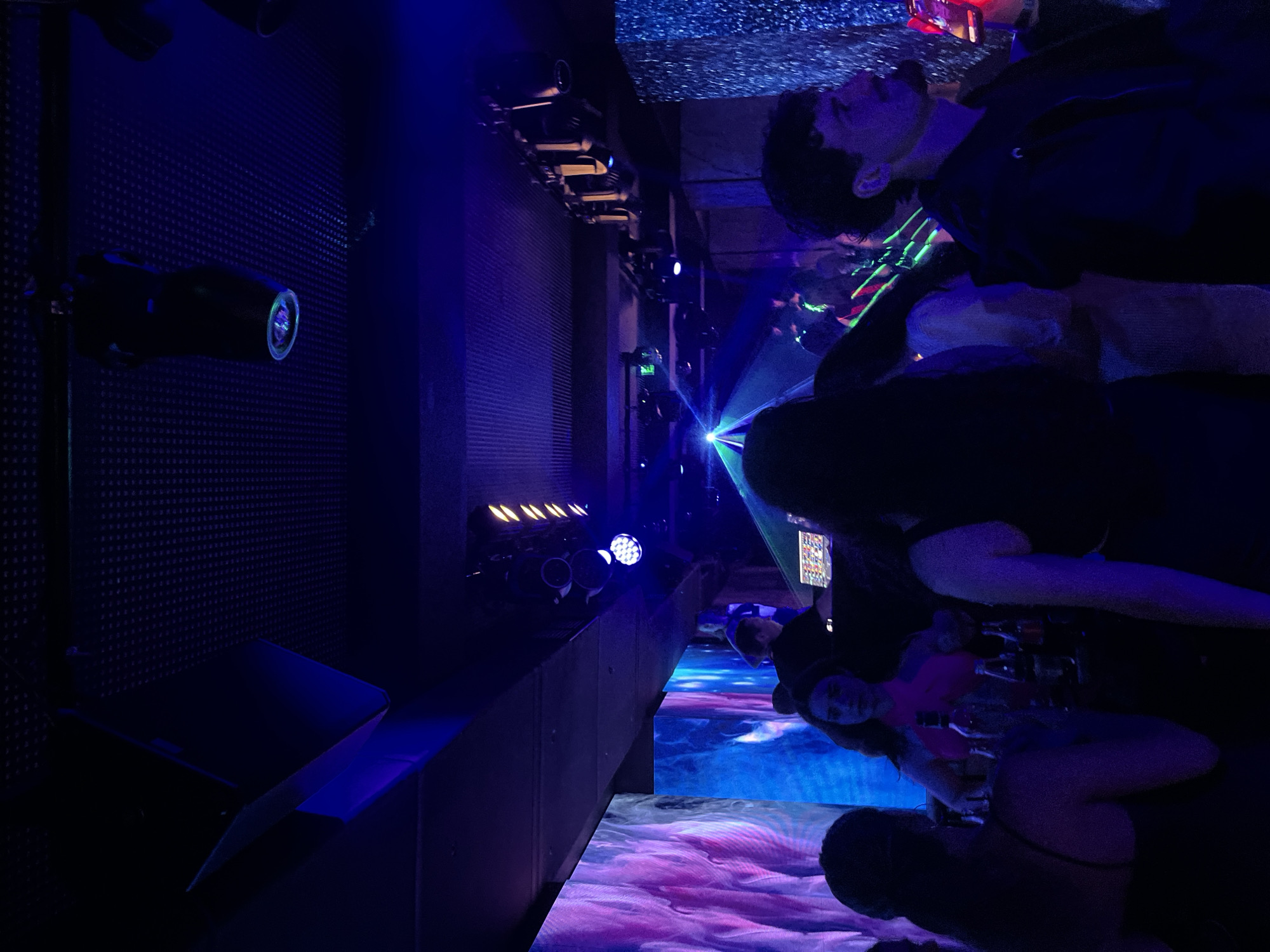 A low-angle, full shot captures a dimly lit nightclub in Sozopol, Bulgaria. The ceiling features a perforated dark paneling with several spotlights and lighting fixtures, some projecting beams of light. The walls are adorned with large LED screens displaying vibrant, abstract, purple and blue patterns on the left, and a glittering, textured silver surface on the right. In the mid-ground, a DJ booth is visible with a person operating equipment, and several bottles of alcohol are arranged on a counter. A crowd of people is present, some silhouetted against the lighting, while others are more clearly illuminated. In the foreground, to the left, a woman with dark hair is seen from behind, wearing a black dress. To the right, a man with dark, curly hair and a stubble is visible in profile, wearing a dark hooded jacket. The atmosphere suggests a lively evening event with music and dancing.
