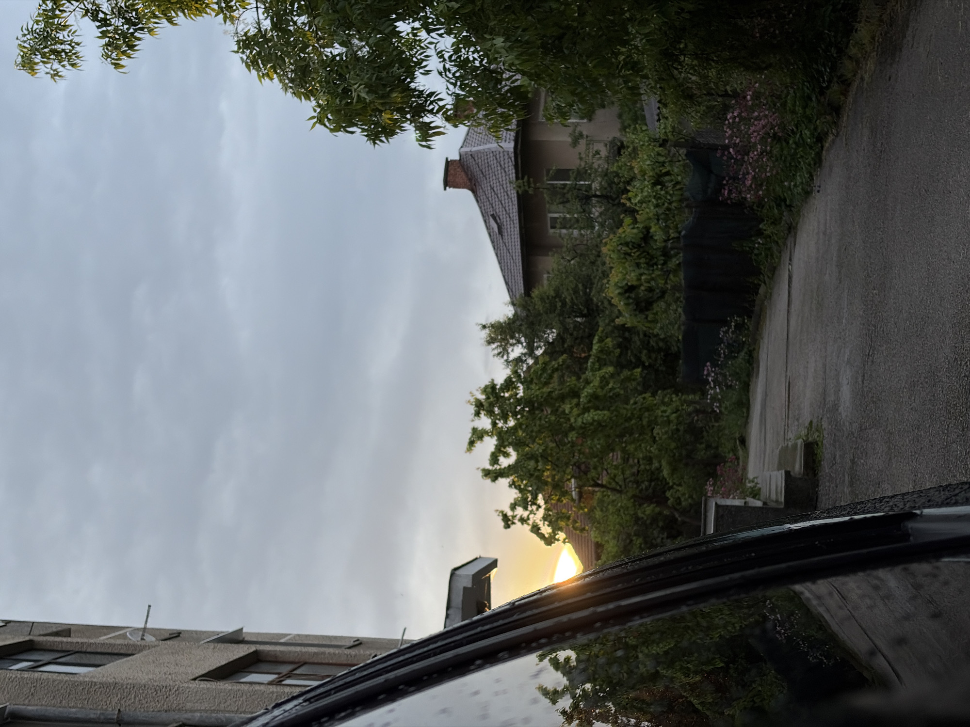 The image shows a street in Botevgrad, Bulgaria, viewed from a low angle, likely from inside a car. The sky is overcast. On the left is a multi-story building with windows. Straight ahead, a residential street curves to the right, bordered by greenery and houses. The edge of the car window, with water droplets on it, is visible in the foreground. The sun is breaking through the clouds.