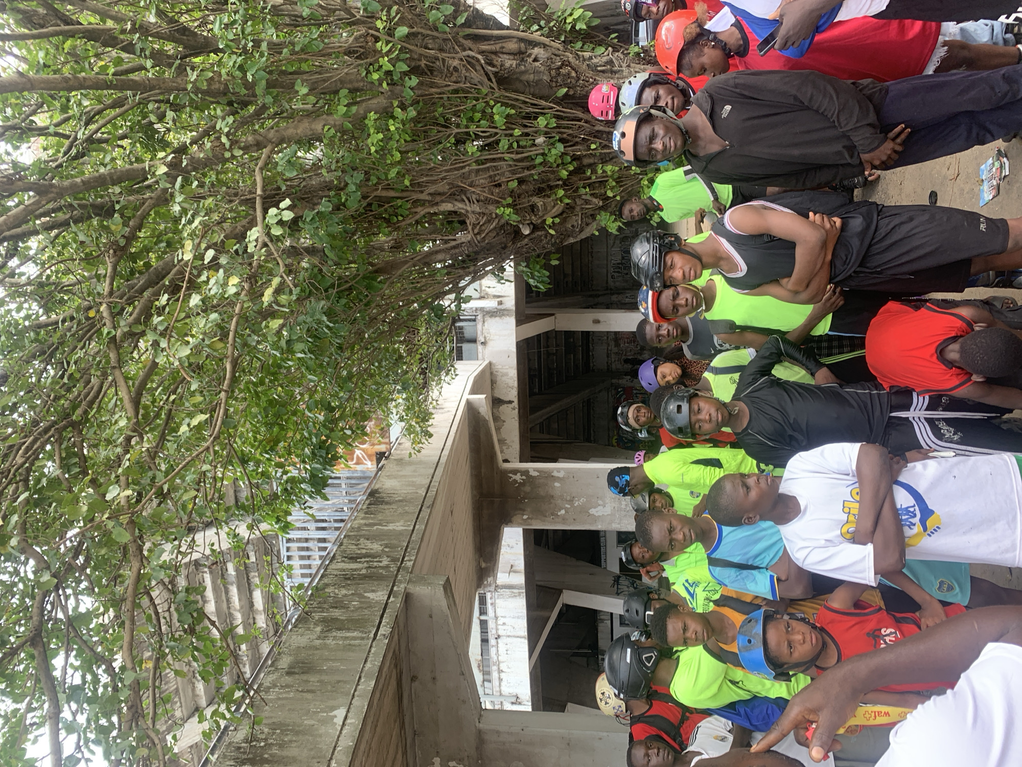 A group of young people, mostly teenagers, are gathered outdoors under a large tree and an overpass structure. Many are wearing helmets, suggesting they may be involved in cycling or a similar activity. The environment appears to be urban, with concrete structures and graffiti visible. The people are dressed in casual clothing, with several wearing bright neon green shirts. Some are looking towards the camera, while others are engaged in conversation or observing their surroundings. The overall scene suggests a community gathering or an organized event taking place in a public space. The presence of a large tree and the overpass structure indicates an outdoor location within a city.