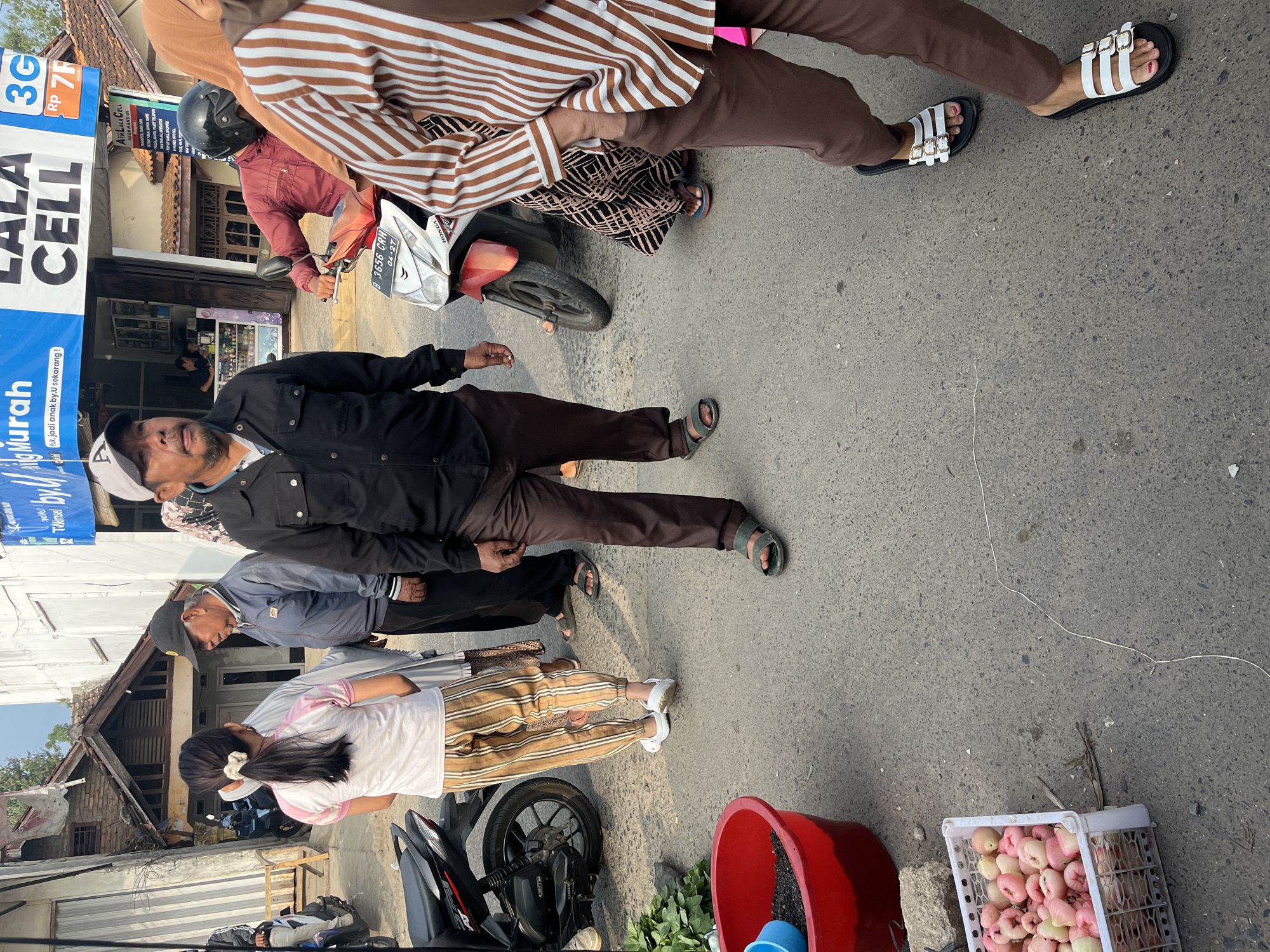 In Kemuning, Indonesia, this street scene captures a vibrant moment of daily life. A man in a black jacket and brown pants stands prominently in the foreground, looking towards the left. He wears a black baseball cap and appears to be speaking. To his right, a person wearing a brown and white striped shirt and brown pants walks away from the camera, their right hand in their pocket. Behind them, a scooter is parked, ridden by a person in a red jacket. In the background, a shop with a blue sign that reads "LALA CELL" is visible. To the far left, a young woman in a white and pink shirt and striped pants faces another person, who is wearing a dark jacket and a headscarf. A red bucket filled with dark soil is to the left of the woman, and a white crate filled with pink rose apples sits on the ground near the bottom left corner. The ground is paved with asphalt, and the overall lighting suggests it is daytime.