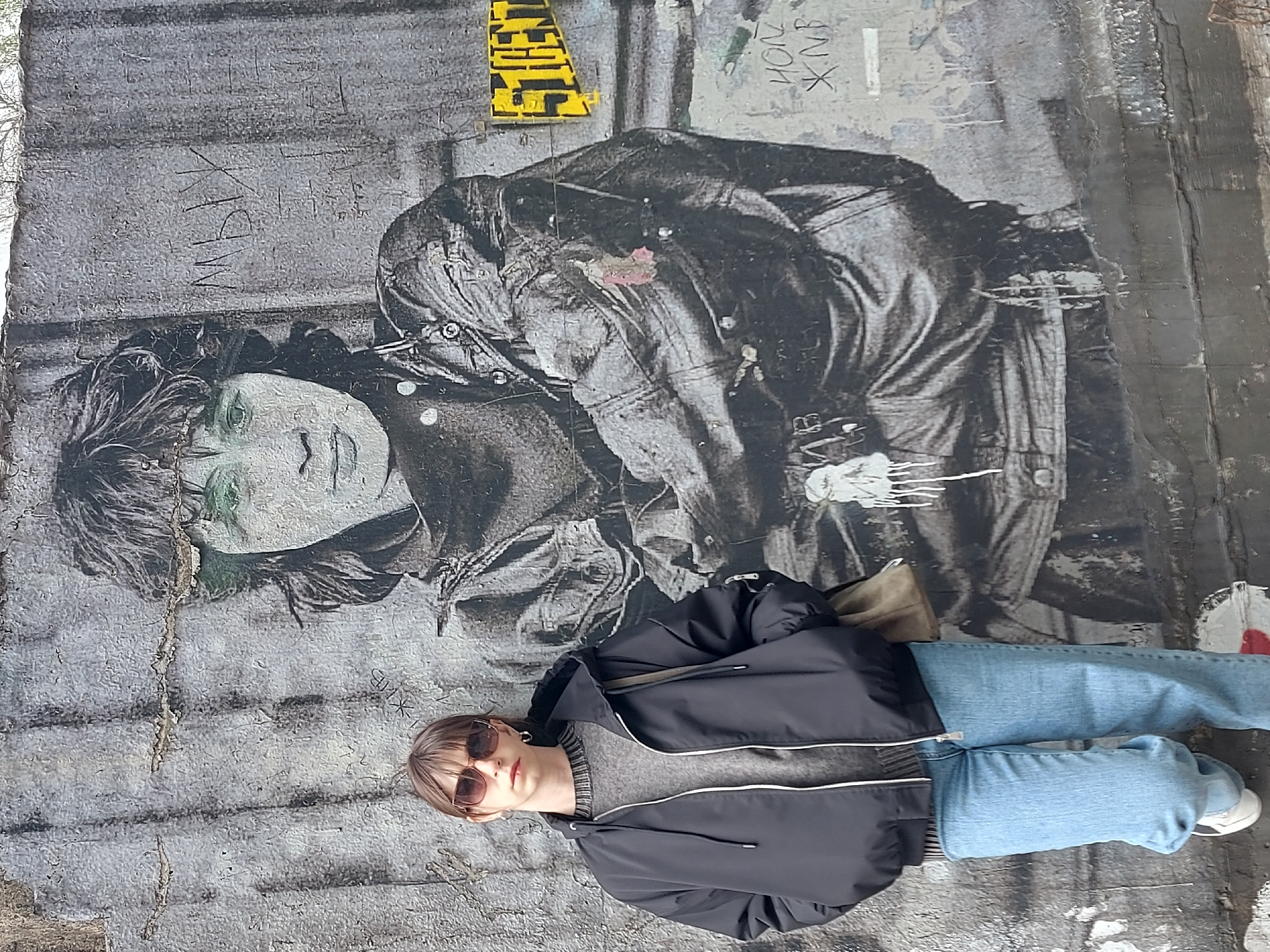 A person lies on the ground next to a mural depicting Jim Morrison in St. Petersburg, Russia. The mural is on a gray wall with additional graffiti. The subject wears a dark jacket, gray sweater, blue jeans, and sunglasses. White sneakers are visible on the subject's feet.