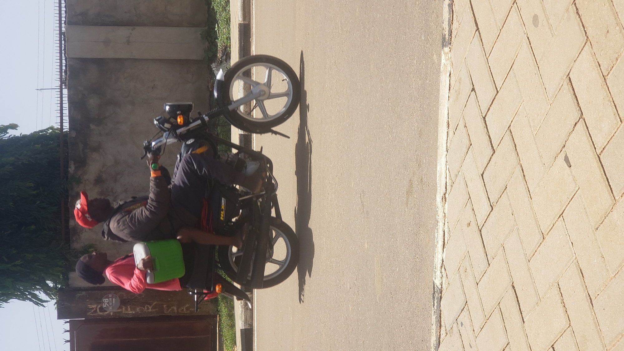 In Narayi, Nigeria, a man wearing a red cap and a dark jacket is sitting on a motorcycle. He is looking to his left, with his right hand holding the motorcycle's handlebars. A young person in a pink long-sleeved shirt and a black headscarf is seated in front of him, also looking to the left. They are holding a green cooler bag between them. The motorcycle is dark-colored with silver wheels. The scene is outdoors on a paved road with a concrete sidewalk to the right. Behind the motorcycle, a grey concrete wall and some green foliage are visible. A metal fence runs along the top of the wall. The sunlight suggests it is daytime, and the shadows indicate it is likely a sunny day. There is no visible text that can be read.