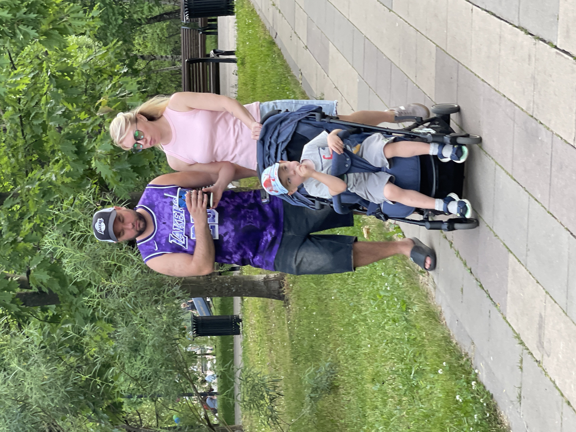 The image depicts a family in a park, likely in Moscow, Russia, given the context provided. A man wearing a purple Lakers jersey and dark denim shorts stands to the left, holding a paper cup. Next to him, a woman with blonde hair pulled back, wearing a pink tank top and denim shorts, pushes a stroller. In the stroller sits a young boy wearing a blue and red baseball cap, a grey t-shirt with a graphic, and grey shorts. The family is on a paved walkway lined with green grass and trees. Benches and trash cans are visible in the background.