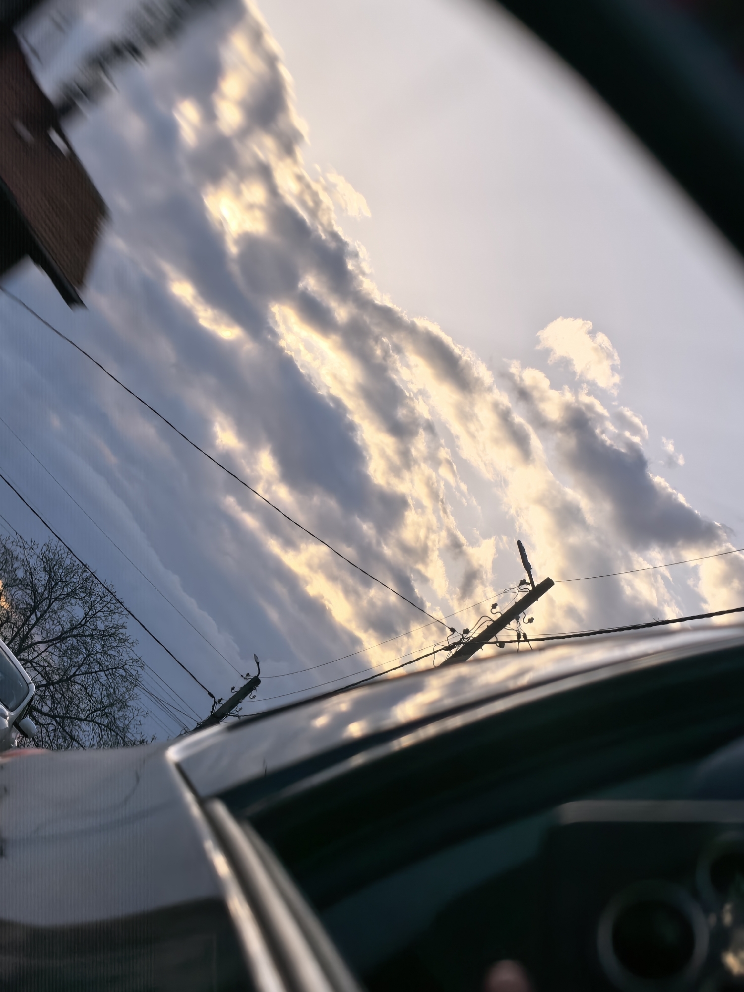 The image shows a view from inside a car, looking up at a sky with striated clouds illuminated by the setting sun. Power lines run across the sky, and a wooden utility pole is visible. A glimpse of a house with a red tile roof is visible on the left, along with a bare tree and a light-colored car. The dashboard of the car is visible in the foreground. The location is a residential area in an unknown city.