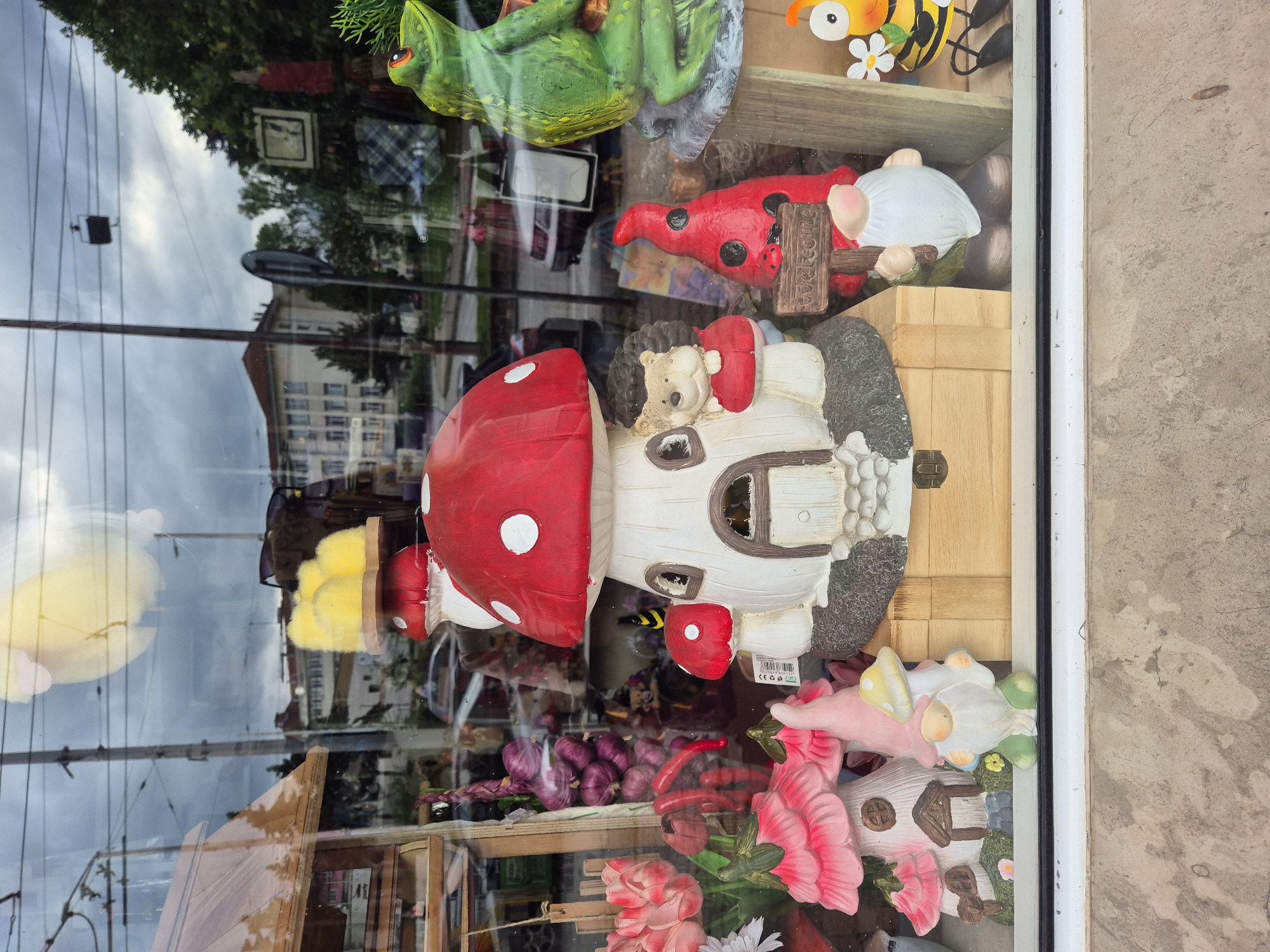 The image displays a shop window filled with decorative garden ornaments, heavily overlaid with reflections from the street outside. The central item in the display is a large, whimsical mushroom-shaped house featuring a bright red cap with white spots, inside of which a small hedgehog figure, wearing a red garment, is nestled.
Above the mushroom house, to the left, a glossy green frog figurine with large eyes is visible. To the upper right, a yellow and black bee figure with white wings and a small white flower adds to the theme. Below the bee, a red ladybug-like gnome or figure holds a small wooden sign that clearly reads "Welcome." Further down on the right, clusters of pink artificial flowers are arranged, alongside what appears to be another small fairy house or decorative structure. In the lower left part of the display, bunches of purple, possibly artificial, garlic or onions are arranged.
The window's surface reflects a cloudy, overcast sky, power lines, and a tall street light pole. Across the street, the reflection reveals a multi-story building with numerous windows, suggesting an urban or suburban setting. Trees and other foliage are also discernible in the reflections. The photograph is taken at an oblique angle, and the rough concrete wall of the building where the shop is located is visible along the far right edge of the frame. A barcode sticker with a "CE" mark is visible on an item near the base of the mushroom house. The scene depicts a daytime setting with diffused light due to the cloudy weather.