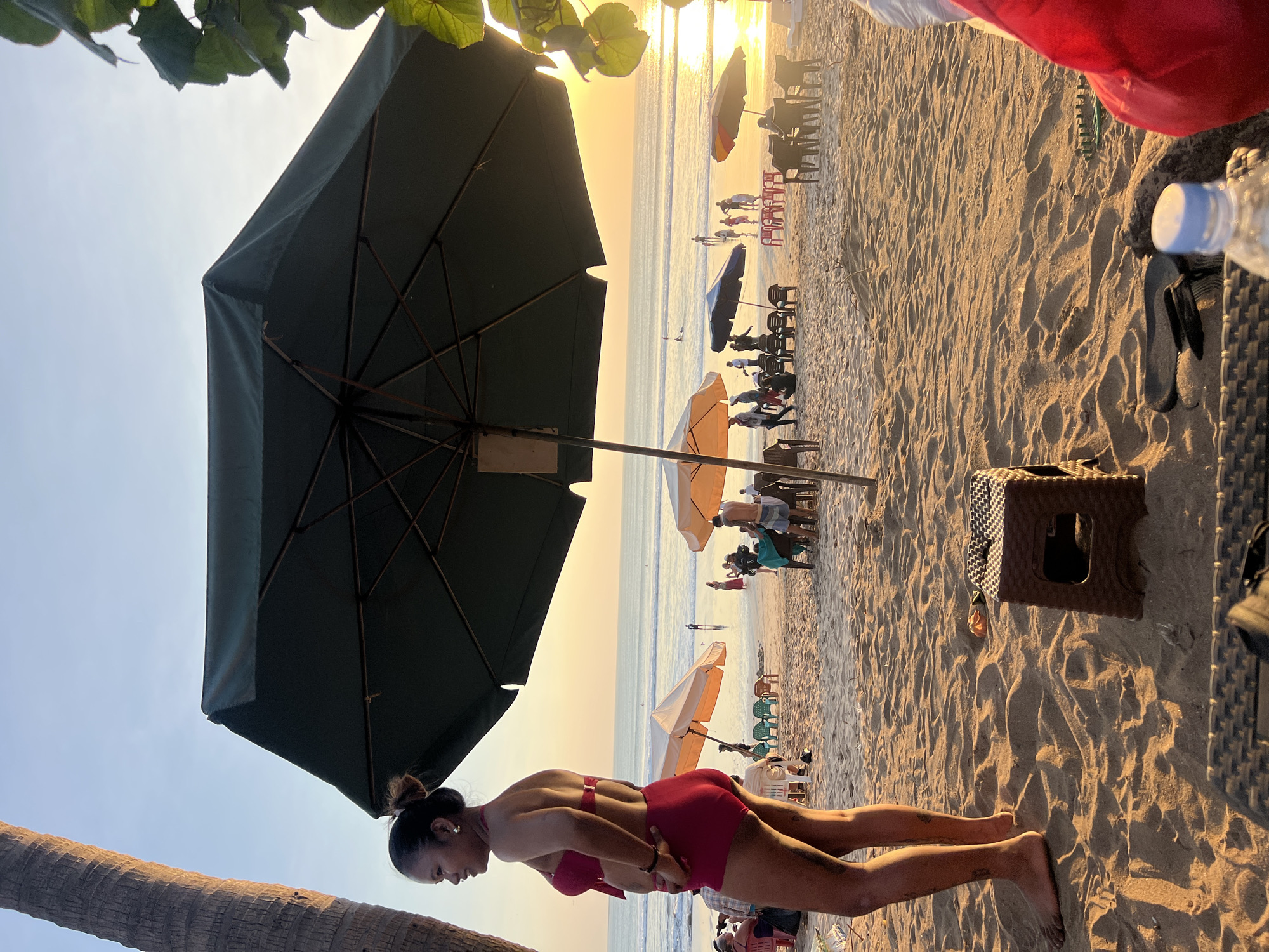A woman in a red bikini stands on a sandy beach in Kuta, Indonesia, facing left and looking towards the ocean. The sun is setting, casting a warm glow over the scene and creating a long shadow of the woman on the sand. In the foreground, a large dark umbrella provides shade. Beyond the woman and the umbrella, beach chairs and other umbrellas are visible, with people scattered throughout the scene enjoying the beach. The ocean stretches to the horizon, with gentle waves and a few figures in the water. The sky is clear with a warm gradient from blue to orange.