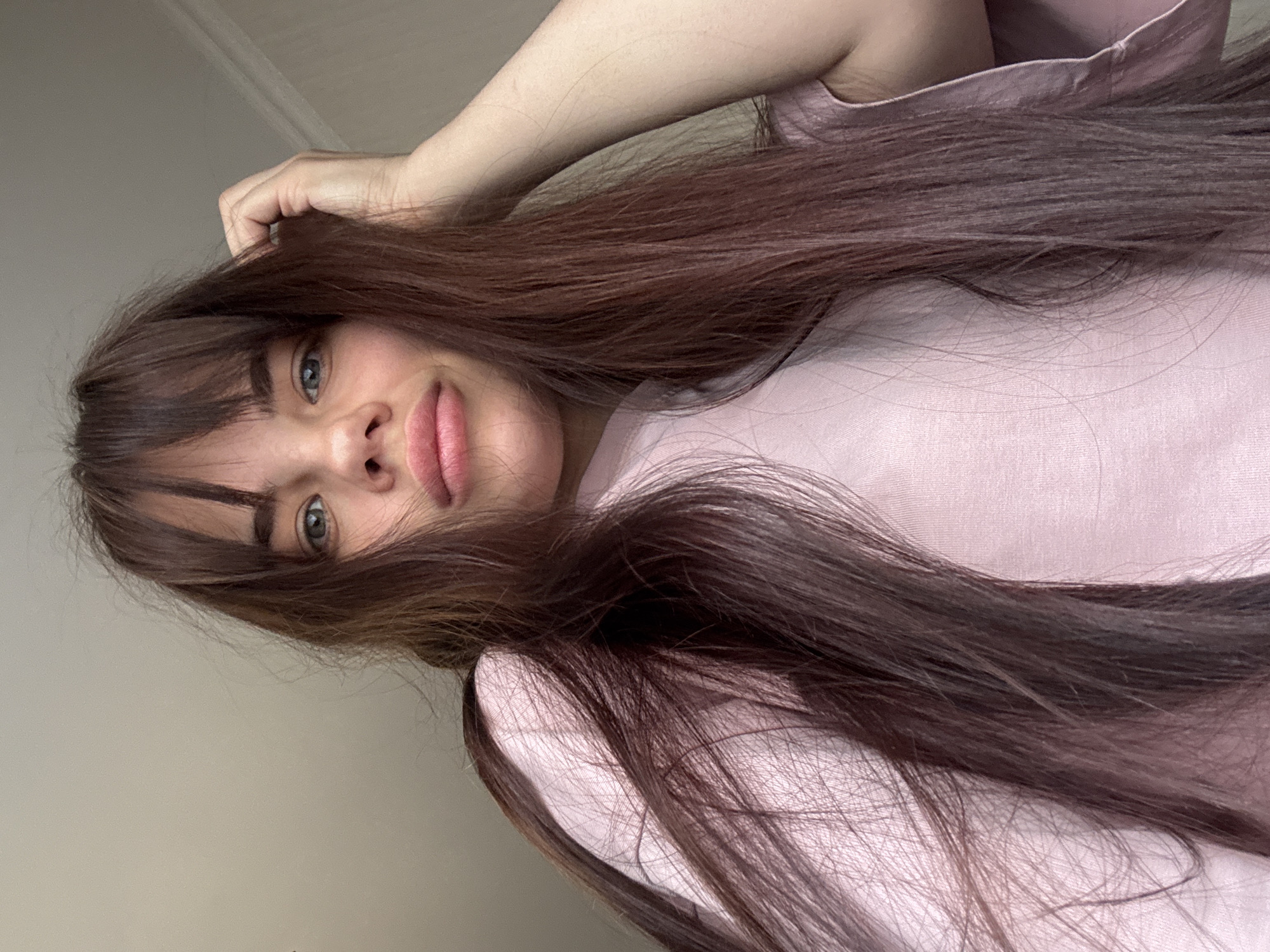 A close-up, inverted portrait features a young woman with long, dark brown hair and light blue-grey eyes, looking directly at the camera. Her face is framed by a fringe and her voluminous hair, which cascades around her shoulders and chest. Her lips are slightly parted. She is wearing a light pink, possibly cotton, top. Her right arm is bent, with her hand resting on the top of her head or in her hair. The background is a plain, light-colored wall, suggesting an indoor setting. The lighting is soft and even, indicating daytime. Her expression is neutral and direct, implying a casual selfie or portrait shot. There are no discernible objects or visible text within the image.
