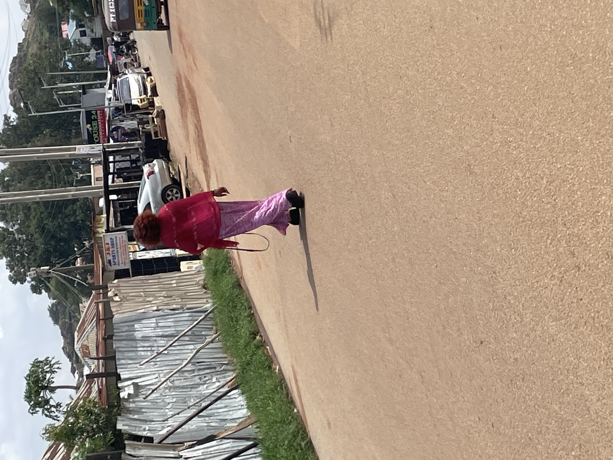 The image shows a street scene in Jos, Nigeria. A person wearing a bright pink jacket and a long pink patterned skirt walks away from the camera on a dirt road. The person is wearing dark shoes and has reddish-brown hair. To the left of the road, a corrugated metal fence separates the road from a row of buildings with rust-colored tin roofs. Some greenery grows along the fence. In the background, there are more buildings, some with white walls and dark roofs. There are also several vehicles on the road, including a silver car, a white SUV, and a green and yellow auto-rickshaw. A sign with "OUSE 24 ACCOMMODATION" is visible on one of the buildings. The sky is partly cloudy, and the overall impression is of a bright, sunny day in an urban area.