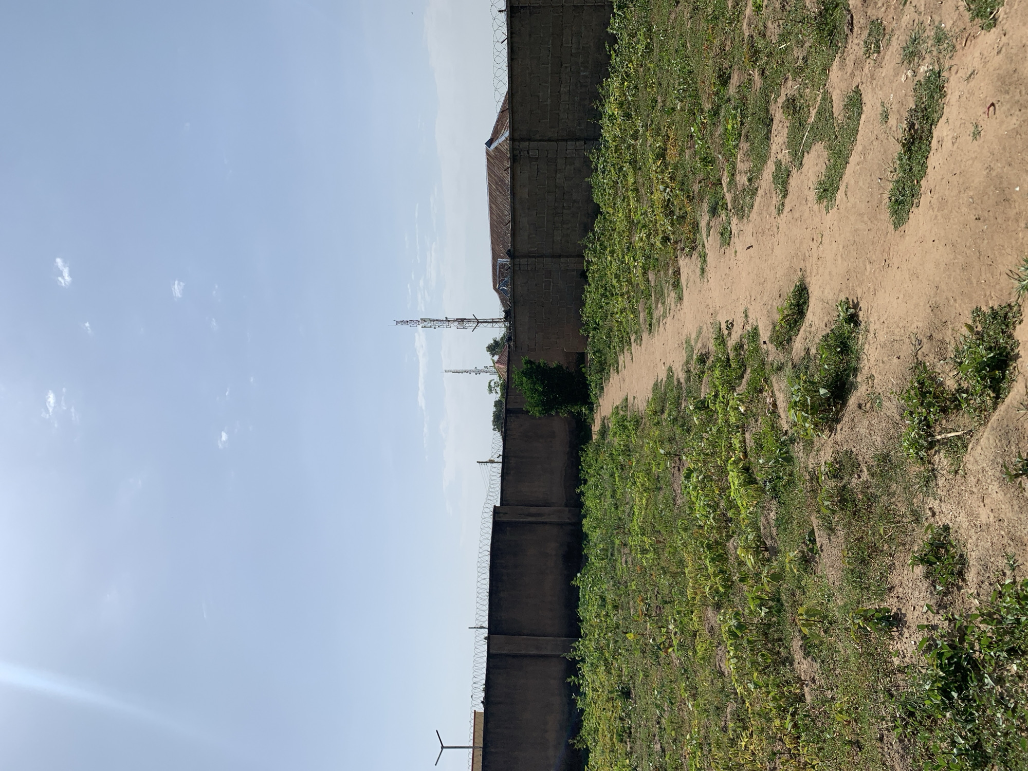 A medium shot depicts an outdoor scene in Rafin Par, Nigeria. The landscape is dominated by a dirt path extending into the distance. To the right of the path, vegetation blankets the ground. To the left, a dark, tall wall topped with barbed wire runs parallel to the path, providing security to a building. Beyond the wall, a telecommunications tower rises into the clear, light blue sky with light white clouds. The scene is bathed in sunlight.