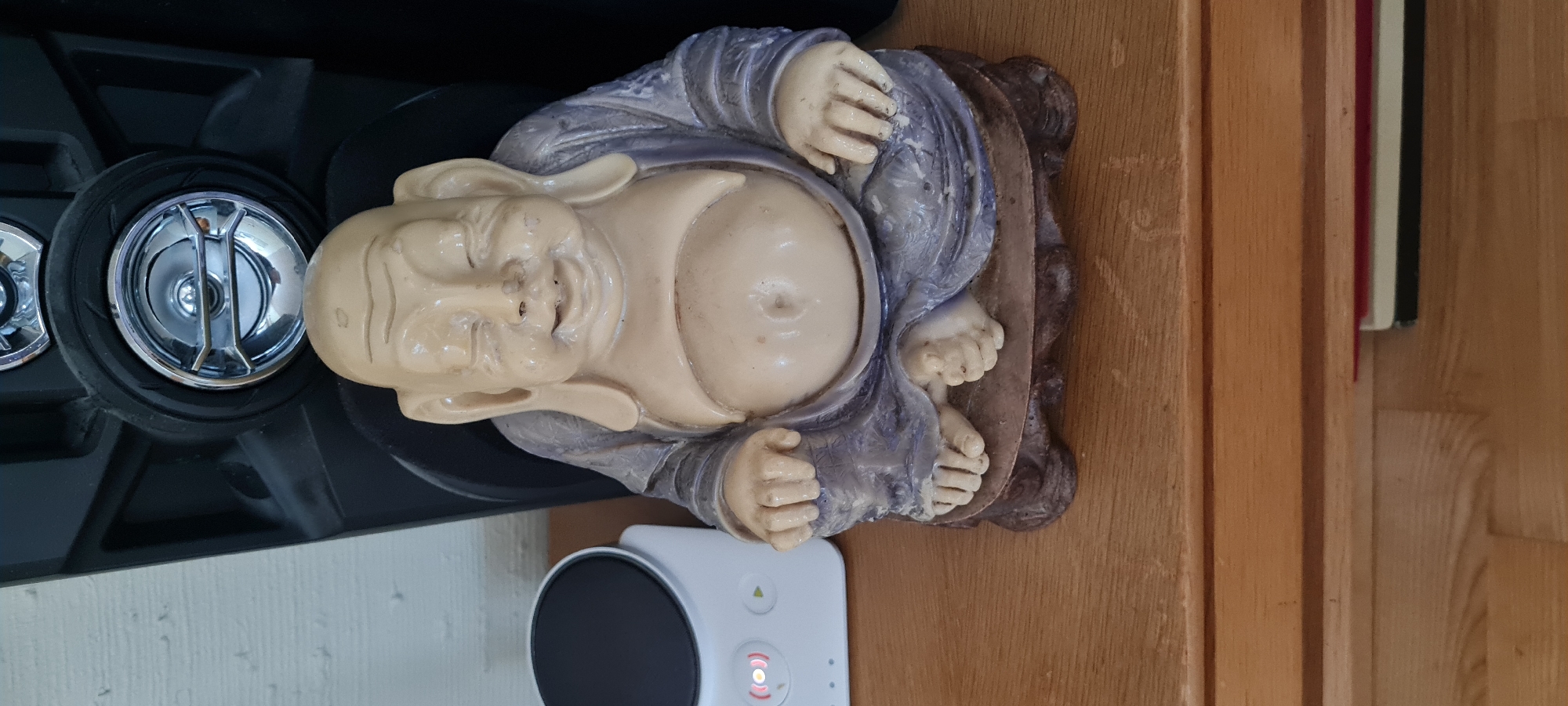 A light-skinned, bald Buddha statue with closed eyes sits on a wooden surface. The statue is wearing a purple robe and has a round belly. To the left of the statue is a white electronic device with a black circular top and red indicator lights. Behind the statue is a large black object, possibly a speaker, with metallic circular accents. The background is a plain, light-colored wall. The floor the statue is on is made of polished wood planks. In the lower right corner, partially obscured, are a few objects that appear to be books or notepads, one with a red edge. The scene appears to be indoors, likely a home or office environment in Borås, Sweden.