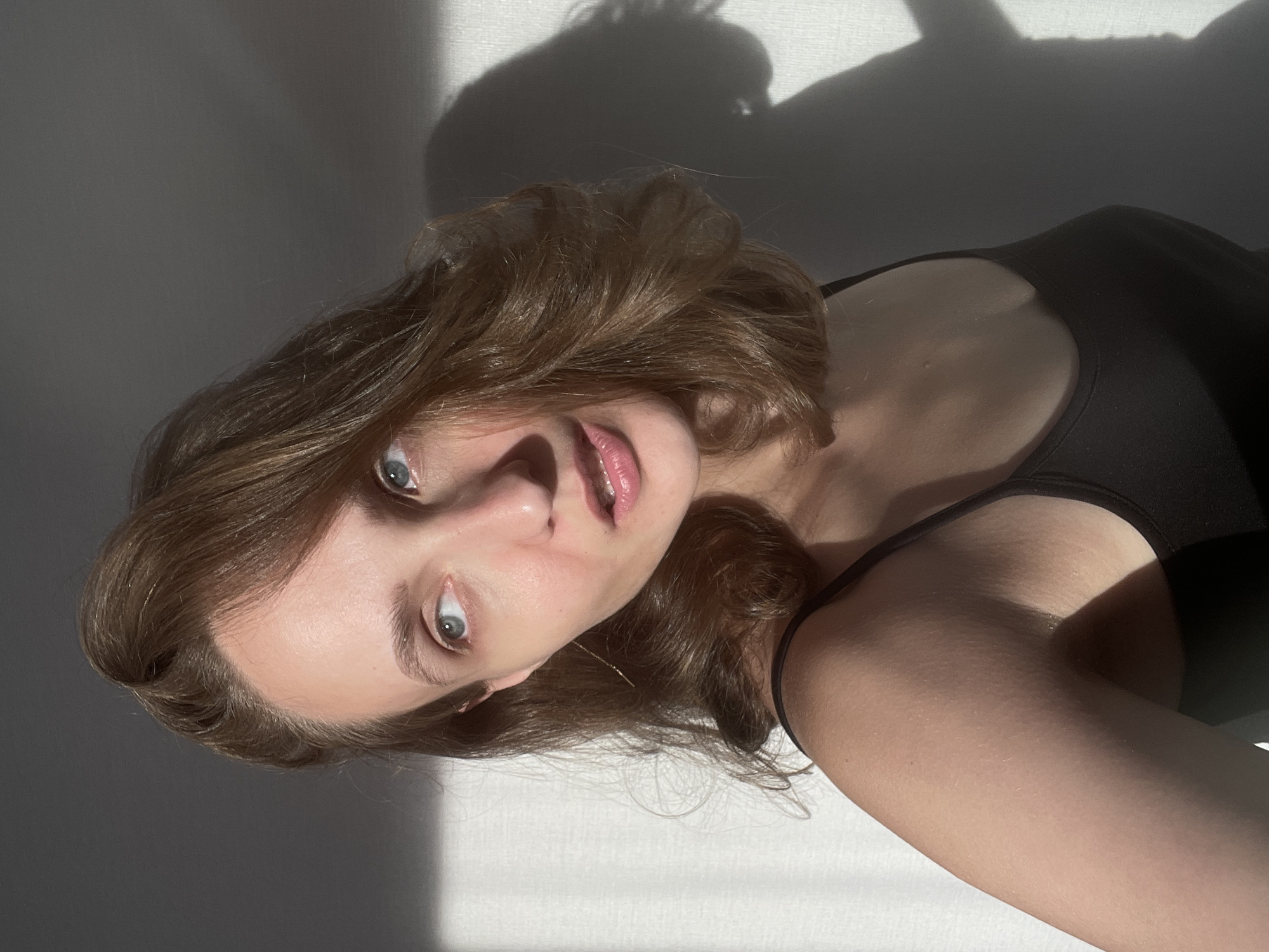 This is a vertical, slightly overexposed, medium shot photograph of a young woman with light brown hair lying on a white surface. She is looking up and to the right of the camera, with a slightly open-mouthed expression. Her eyes are light blue and her lips are pink. She is wearing a black tank top.
The lighting suggests it is daytime, with strong sunlight casting shadows on the wall behind her. The shadows are abstract and form interesting shapes. There is no visible text in the image. The setting appears to be indoors, possibly a bedroom or studio, with a plain white surface and a textured wall. The woman's pose and facial expression convey a relaxed and perhaps playful mood. The location context provided is Подшивалово, Russia, but there are no specific visual cues in the image to confirm this location.