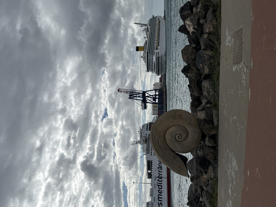 The image presents a daytime scene at the port of Puerto del Rosario, Fuerteventura, Spain, under an overcast sky. In the immediate foreground, a paved promenade, featuring reddish-brown asphalt on the left and grey concrete with an embedded metal grate on the right, extends across the frame. Directly beyond the promenade, a line of dark, irregular rocks forms a boundary. Centrally positioned on these rocks is a large, light-toned sculpture of a spiral shell, oriented horizontally. The midground features the calm, grey-blue water of the harbor. On the left, a large white ferry, partially displaying "osmediterrán" in red lettering on its hull, is visible, possibly docked. Adjacent to the ferry, a prominent dark blue port crane with a red apex stands, surrounded by stacked shipping containers on the dock. To the right, a large white cruise ship, identifiable by its yellow funnel, is either docked or at anchor. The background consists entirely of a dense, grey cloud cover, with minimal patches of brighter sky. No individuals are present in the scene.