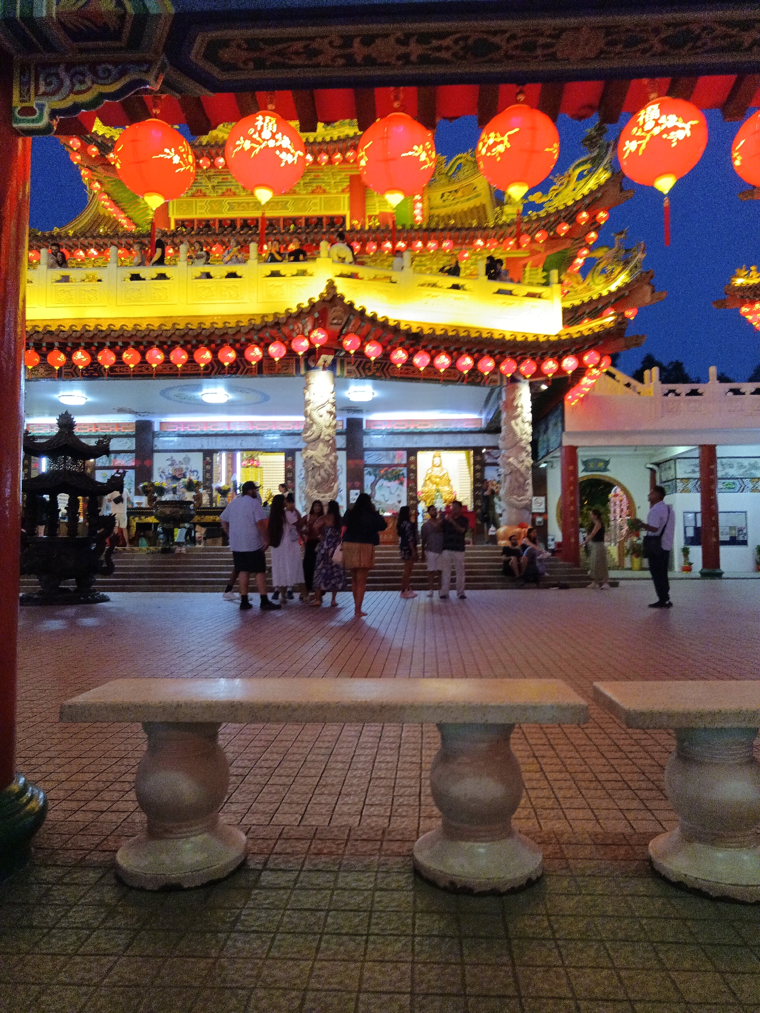 This image depicts a view of Thean Hou Temple in Kuala Lumpur, Malaysia, at dusk. The temple's ornate, multi-tiered roofline is illuminated by a bright yellow light, and adorned with numerous red lanterns. Some of these lanterns hang prominently in the foreground, featuring intricate floral cutouts. Below the main temple structure, a paved courtyard area is visible, with a line of red lanterns strung across. In the courtyard, several people are present, some standing and conversing, others seated. A large, dark, intricately designed incense burner stands to the left. In the immediate foreground, two stone benches with decorative bases are positioned on the tiled courtyard floor. The sky above is a deep blue, indicating the transition from day to night.