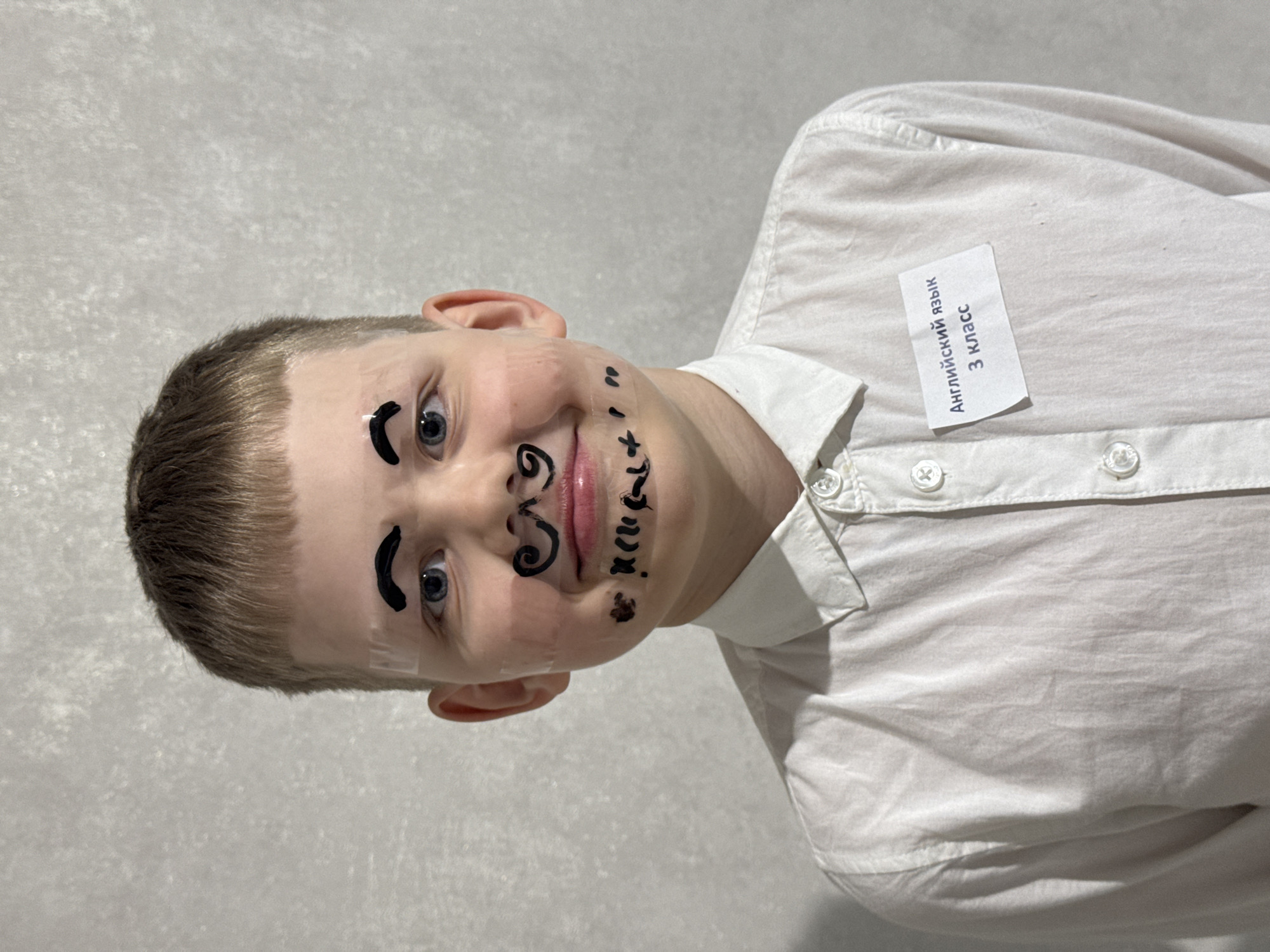 A young boy is centered in the image, his face decorated with drawn-on eyebrows, a mustache, and other markings in black. He wears a white button-down shirt with a name tag that reads "Английский язык 3 класс" (English language 3rd grade) attached to the collar. The background is a plain, light-colored wall. The location appears to be inside a building, potentially a school or home. The image suggests a playful or educational setting. The scene is likely located in Russia, as the name tag is written in Russian. No indication as to the city Gornyi can be seen.