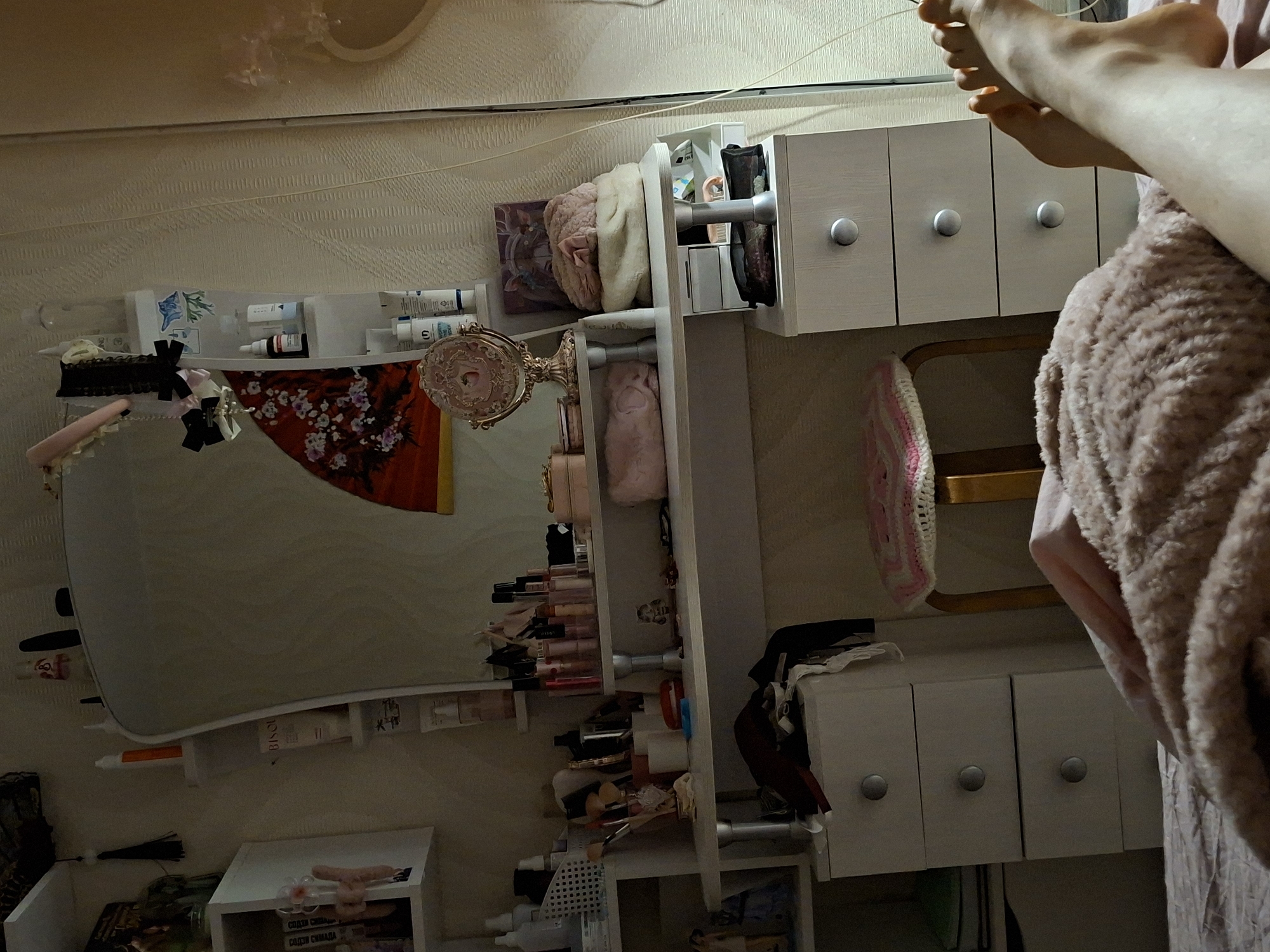 The image, taken in Chaikovsky, Russia, depicts an indoor scene, likely a bedroom or dressing area, viewed from a slightly high angle and rotated 90 degrees counter-clockwise.
The primary subject is a person whose bare feet and lower legs are visible in the upper right portion of the frame (if oriented correctly), resting on a soft, light-pink, textured blanket or garment.
The room is furnished with several white shelves and small cabinets with circular silver knobs, organized around a large, rectangular mirror that occupies the left-center of the image. The mirror reflects parts of the room, including the wall and some objects. To the right of the mirror, an ornate golden-colored table mirror with a pink frame is placed on a shelf.
Numerous objects, predominantly cosmetics and personal care items, are arranged on the shelves. These include various tubes, bottles, brushes, and lipsticks. A small, clear organizer on one shelf holds several items, one with a blue design. Other accessories like black bows, pink headbands, and a small pink crocheted item are also visible. A dark red fabric with a white floral pattern is draped near the mirror. Below the main mirror, a smaller white shelf unit with an open drawer contains books or packages, one of which appears to have text in Cyrillic. On one of the visible tubes, the brand "BISOU" is discernible. Faintly visible text on a package or book on a lower shelf reads "СПАСИБО СИЛАЛА" (SPASIBO SILALA).
The activity taking place is a person relaxing, with their feet elevated. The overall setting suggests a personal and lived-in space, possibly a vanity area where beauty products are stored. The lighting appears to be soft indoor illumination, and the walls are covered with a light-colored, textured wallpaper. A thin light-colored cord runs horizontally across the upper wall. The time of day cannot be precisely determined but does not appear to be bright daylight.