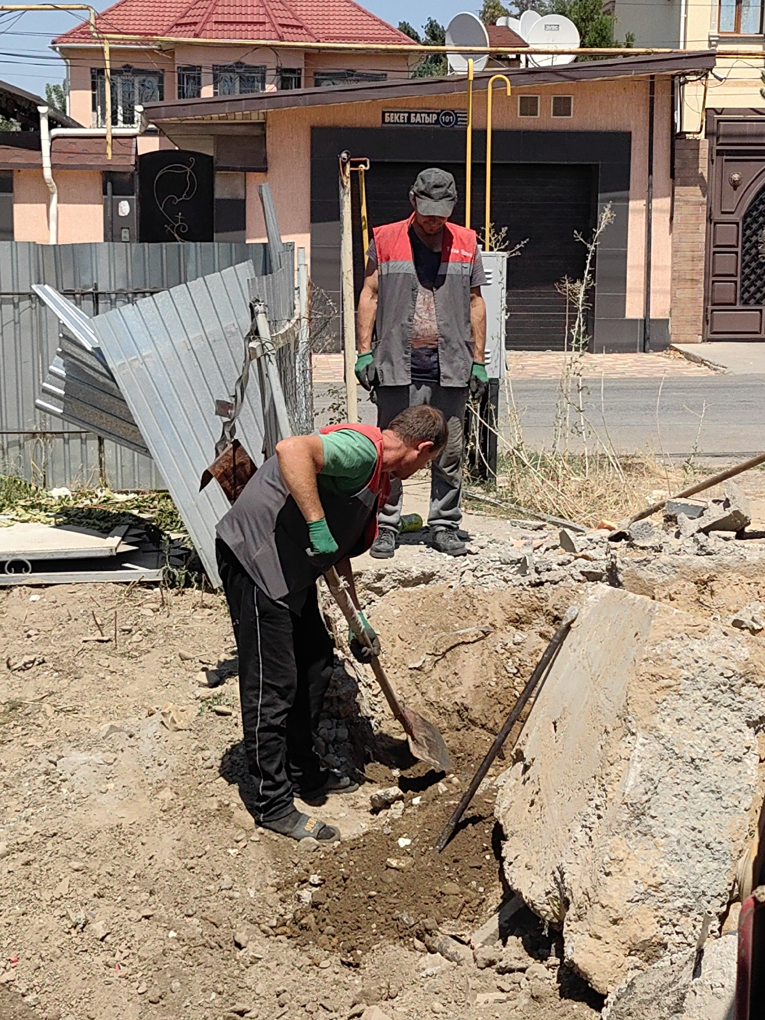 Two construction workers are engaged in digging a trench in a dirt area. The worker in the foreground, wearing a green shirt, grey vest, black pants, and black sandals, is actively digging with a shovel. The second worker, wearing a red vest and dark pants, stands behind him, observing. The scene appears to be in a residential or semi-commercial area, with a building and a metal fence in the background. The building has a pink facade and a red tiled roof, with visible satellite dishes on top. A street sign with Cyrillic text, "БЕКЕТ БАТЫР (101)", is visible on the building, indicating the location as Beket Batyr Street in Shymkent, Kazakhstan. The ground is uneven and filled with loose dirt, rocks, and debris, suggesting ongoing construction or demolition work. A damaged metal sheet leans against the fence. The sky is clear and the lighting suggests it is daytime.