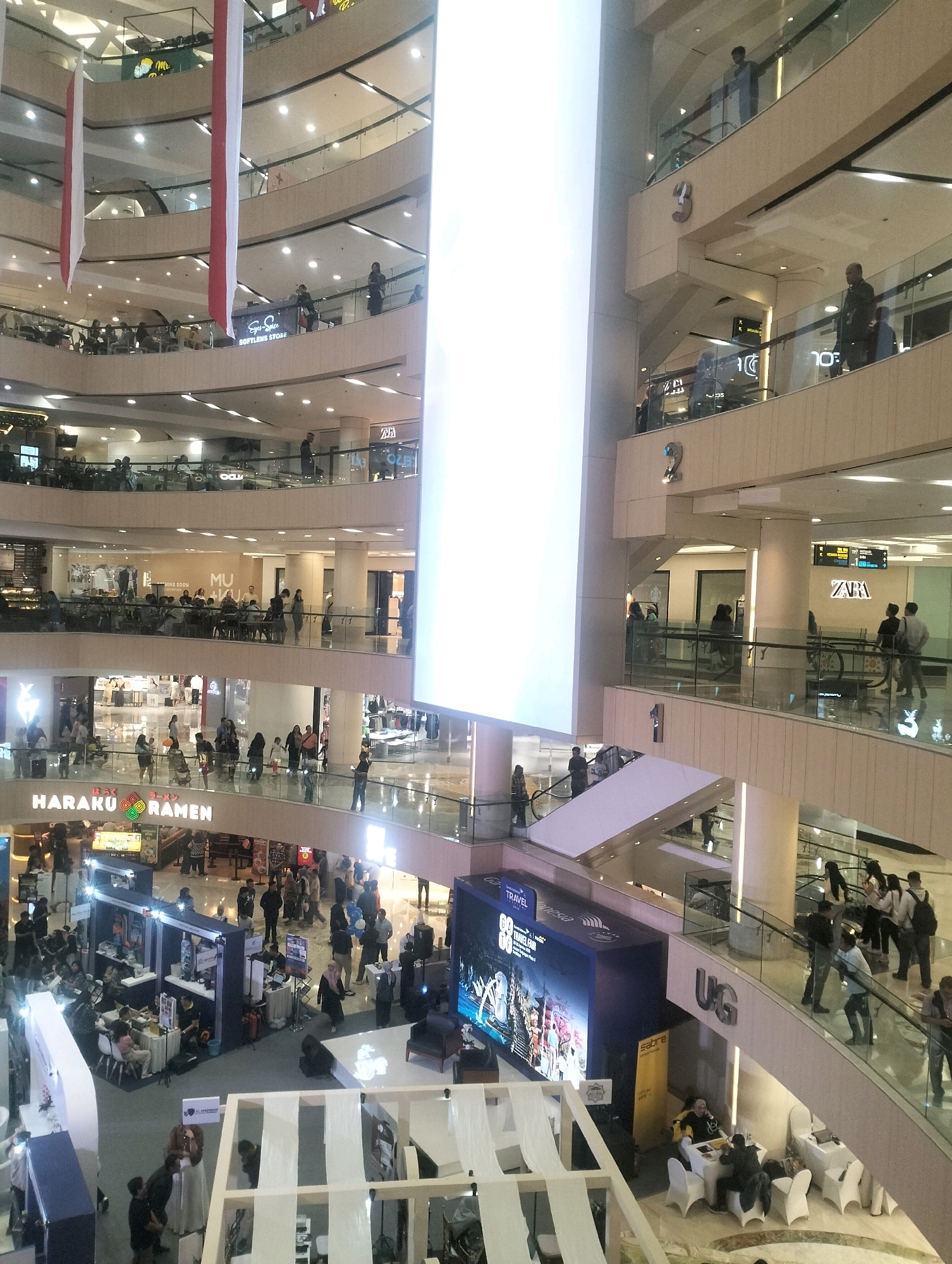This image captures a wide, multi-level interior of a modern shopping mall in Surabaya, Indonesia, likely during a daytime event or busy period. The architecture features cream-colored walls, glass railings, and multiple escalators and staircases connecting the different floors. Several large, brightly lit vertical panels dominate the center of the atrium, one of which is a large screen displaying what appears to be an advertisement or promotional material.
On various levels, there are numerous shops and stalls. Signs visible include "HARAKU RAMEN," "ZARA," and what seems to be an "EYES UP SOFTLENS STORE." People are observed moving between floors via escalators and walking along the walkways. Many are browsing, conversing, or queuing. There is a distinct event setup on the ground floor, with multiple dark-colored booths and tables, suggesting a fair or exhibition. Several individuals are interacting at these booths, and some are seated at tables. Red and white banners, possibly national flags, are hanging from the upper levels, adding a festive or patriotic touch. The mall is illuminated by a combination of overhead lighting and the bright screen, creating a well-lit, expansive atmosphere.